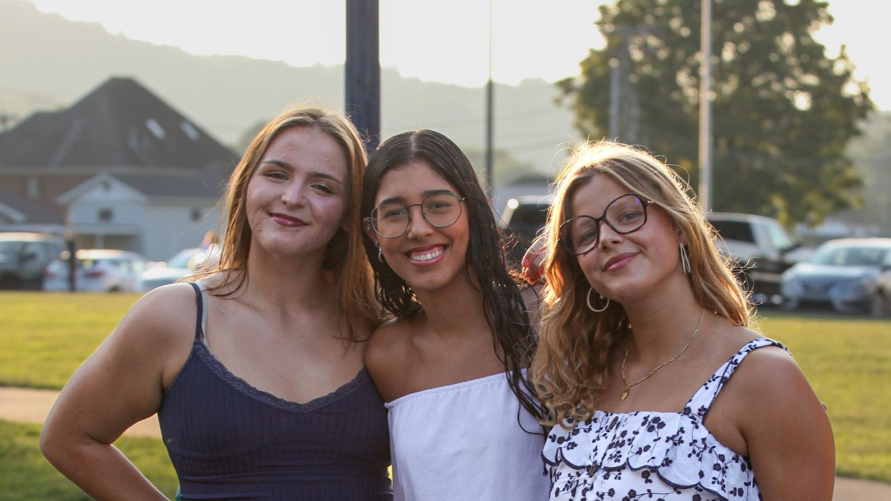 Three female Union Commonwealth University students are posing for a photo in the bright evening sunlight outside the Conway Boatman Chapel. They are standing together, arms around each other, all smiling and dressed in formal attire. The chapel parking lot filled with cars can be seen in the background.