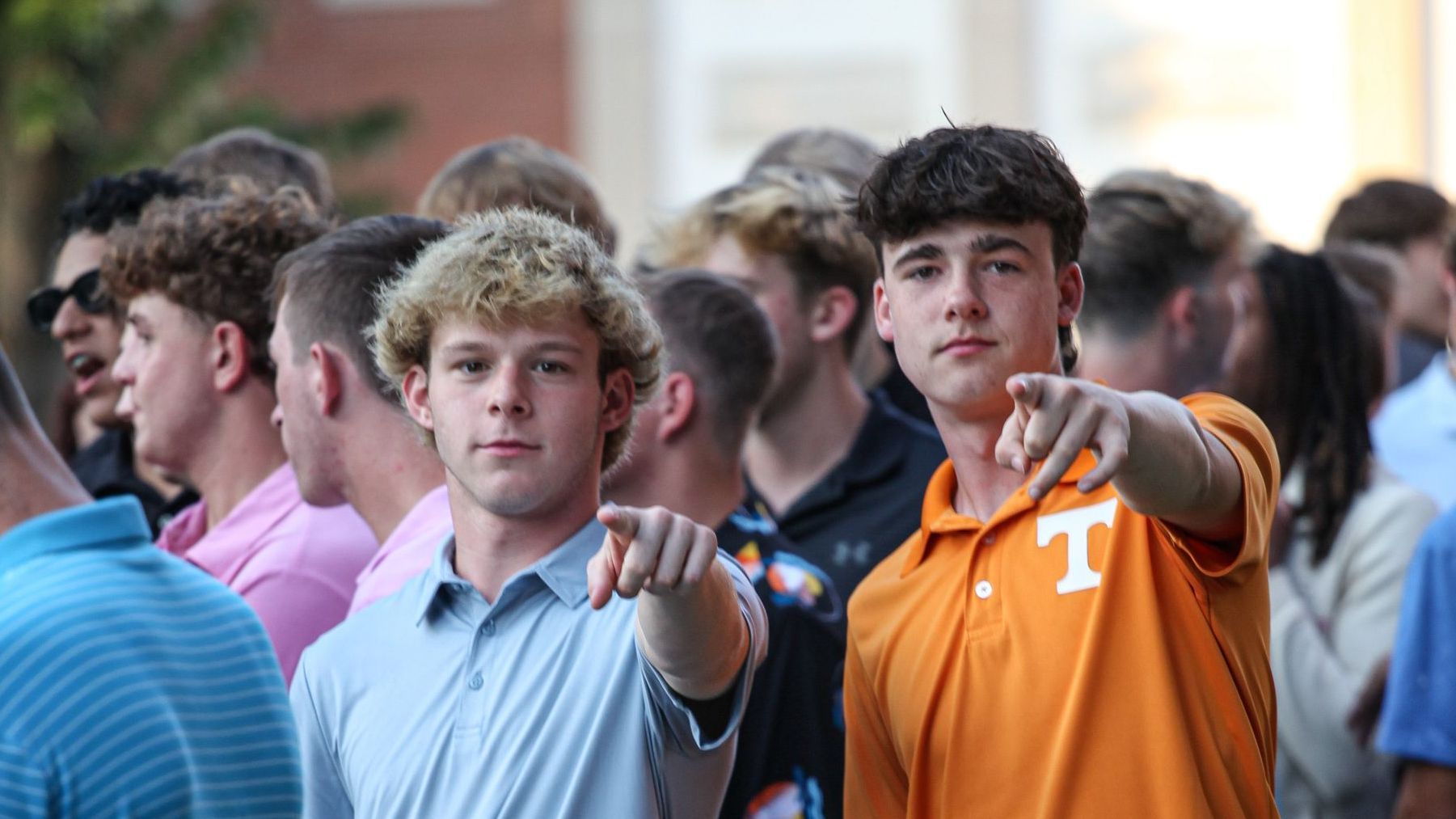 Two male Union Commonwealth University students wearing polo shirts stand pointing at the camera for a photo, outside the Conway Boatman Chapel, among a larger gathering of students, faculty, staff, and community members.