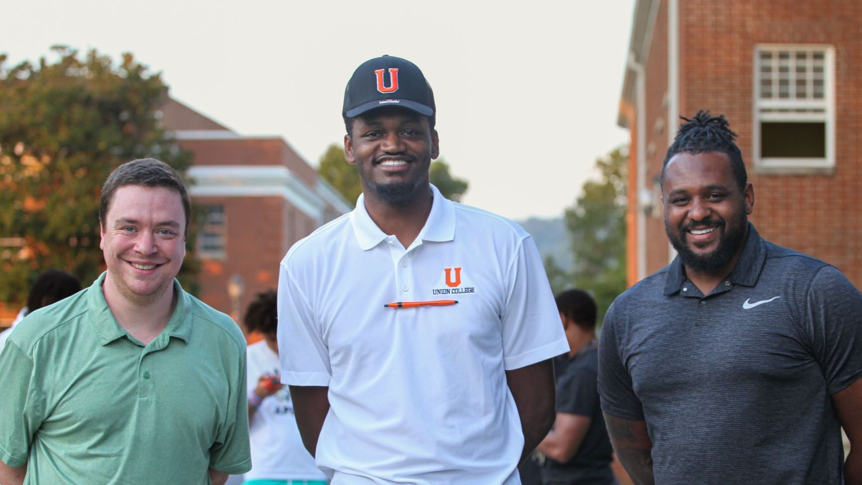 James Becknell, Joshua Falls, and Coach Rodney Haddix stand together outside the Conway Boatman Chapel before Convocation, smiling and dressed in polo shirts. Joshua's shirt bares the Union logo with a pen clipped beneath it. A crowd is visible in the background.