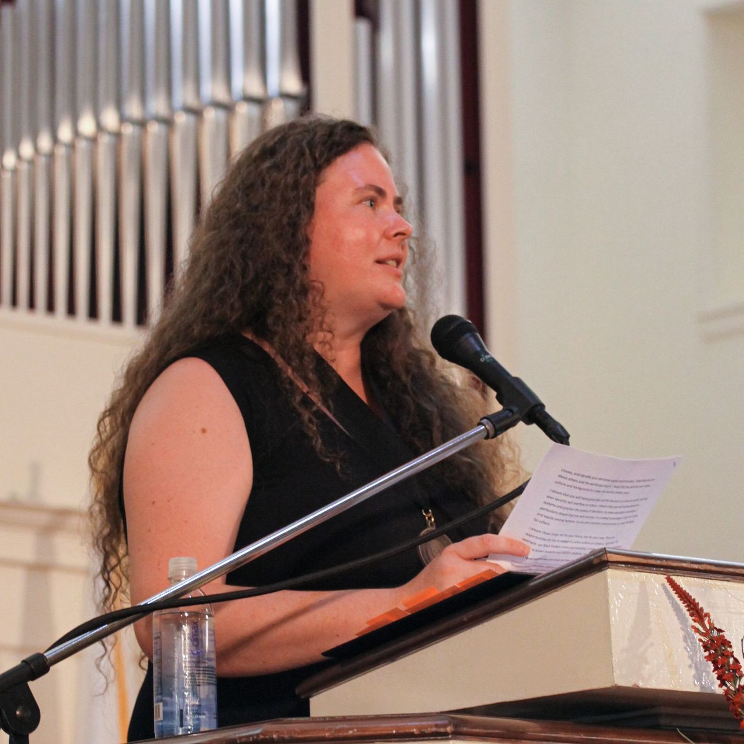 Libby Megna stands at a podium with a mounted microphone in front of the Conway Boatman Chapel delivering her Convocation address. She is wearing a formal black dress and holding what appears to be a copy of her address.