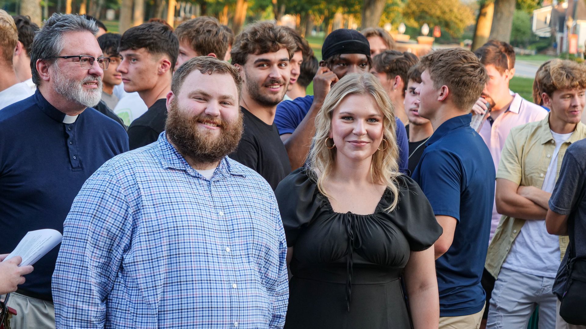 Travis Hensley and Kassidy Koogler stand smiling together among a larger gathering of students, faculty, staff, and community members outside the Conway Boatman Chapel. A mix of casual and business-casual attire can be seen on all present.