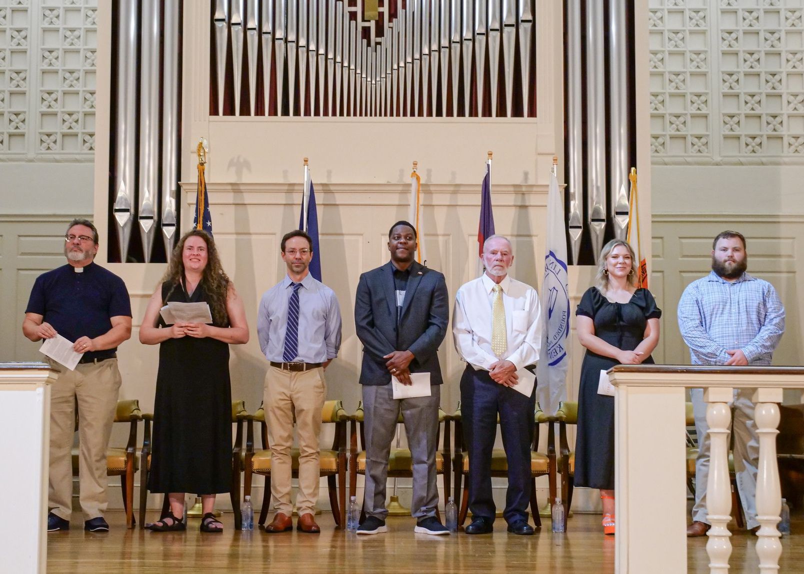Rev. David Miller, Libby Megna, Dr. David Powell, Dr. DJ Washington, Ronnie Moore, Kassidy Koogler, and Travis Hensley stand side-to-side on the Conway Boatman Chapel's stage. They are all dressed in formal attire and holding various papers. Behind them, a collection of flags is mounted beside each person's chair.