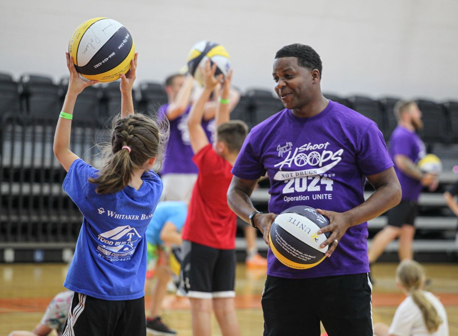 Washington is wearing a purple Shoot Hoops Not Drugs t-shirt while holding a basketball and talking to children holding basketballs up over their heads