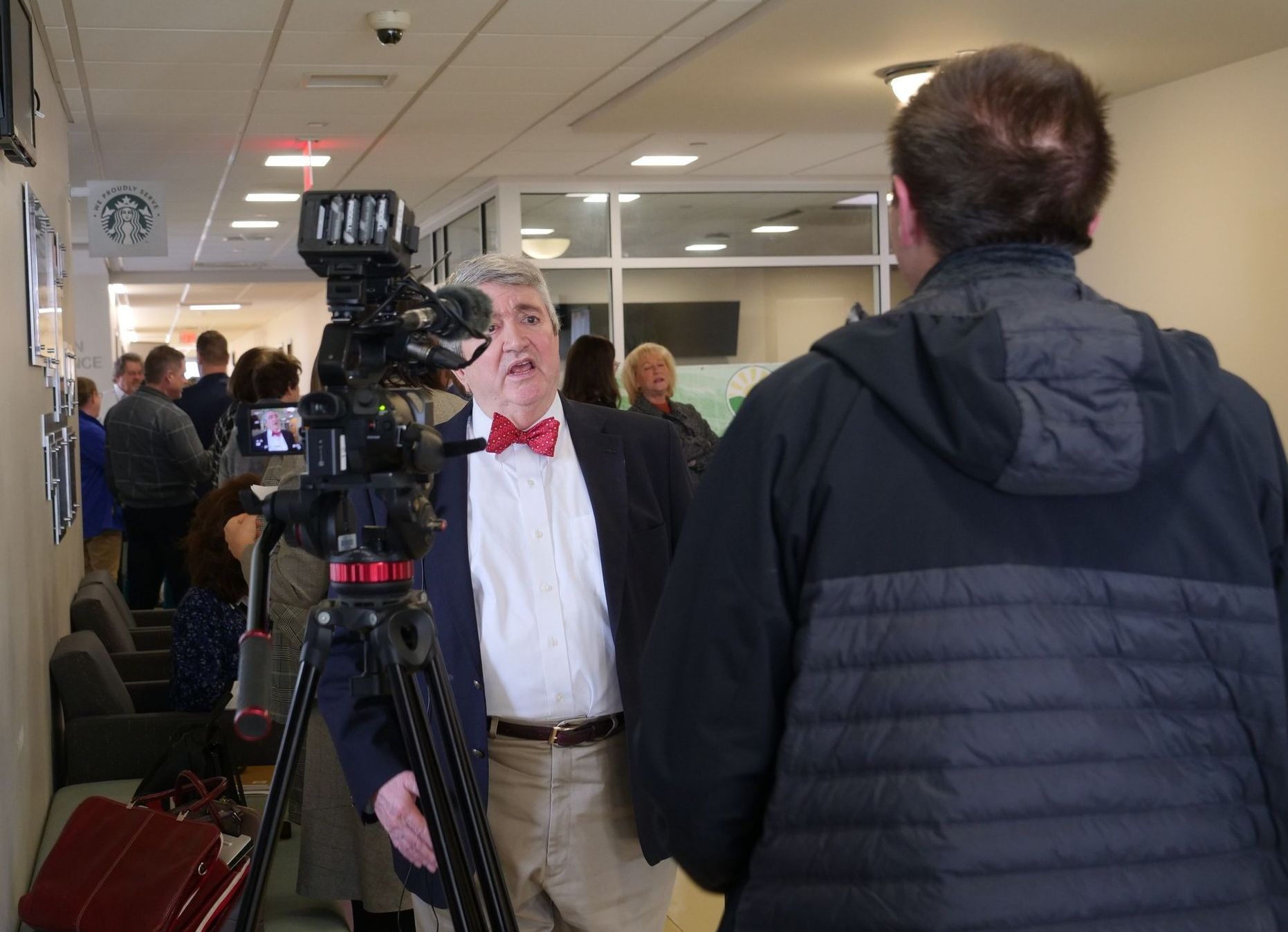 Dr. Al J Mooney talking to the press with a camera recording him in a hall with a crowd of people behind.