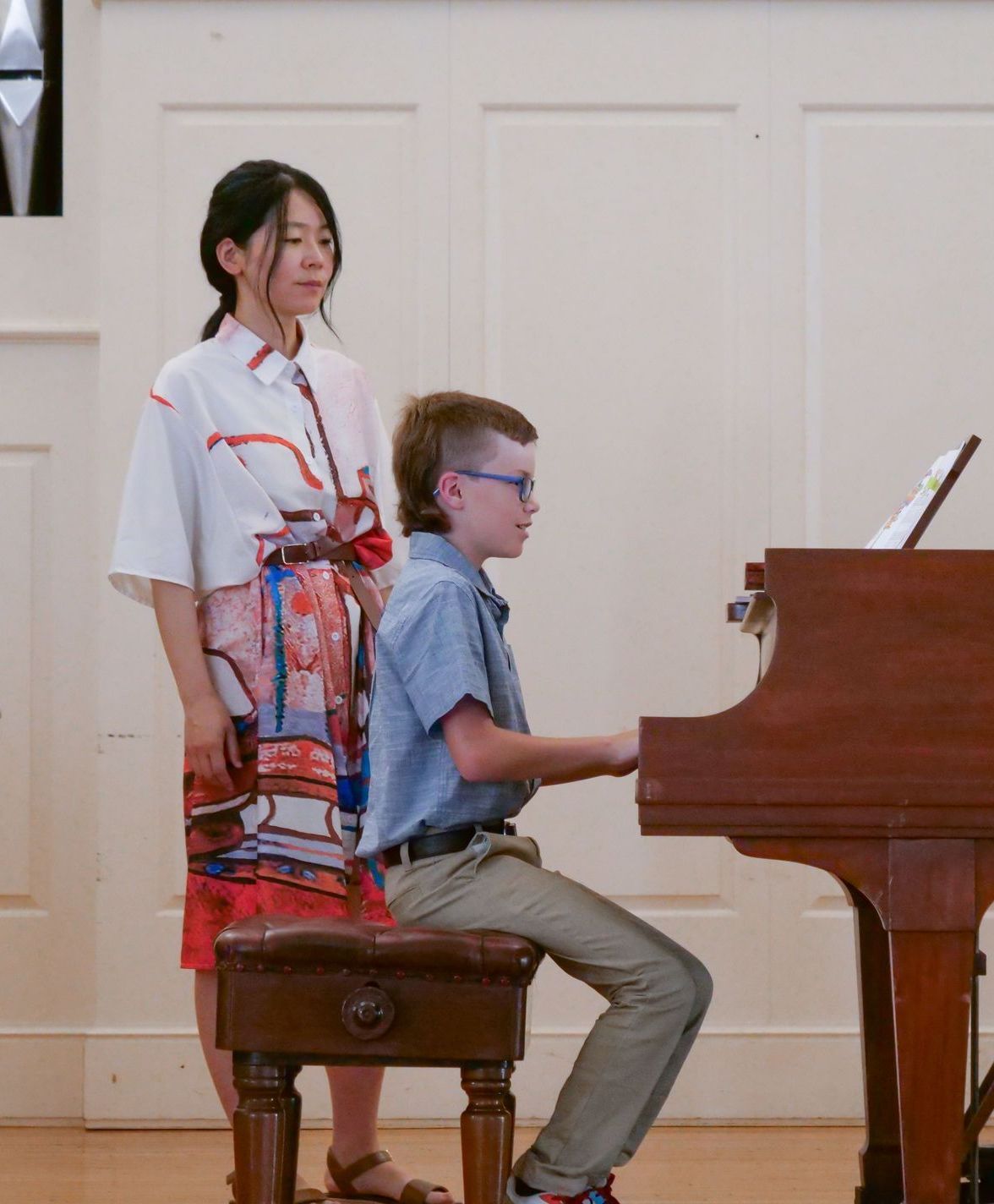 Young David Whitaker sits on a piano bench wearing dress clothes and practicing on the brown wooden piano in Conway Boatman Chapel. Dr. Fujimura stands beside him wearing a vibrant red, white, and blue dress and looking toward the music rack on top of the piano.