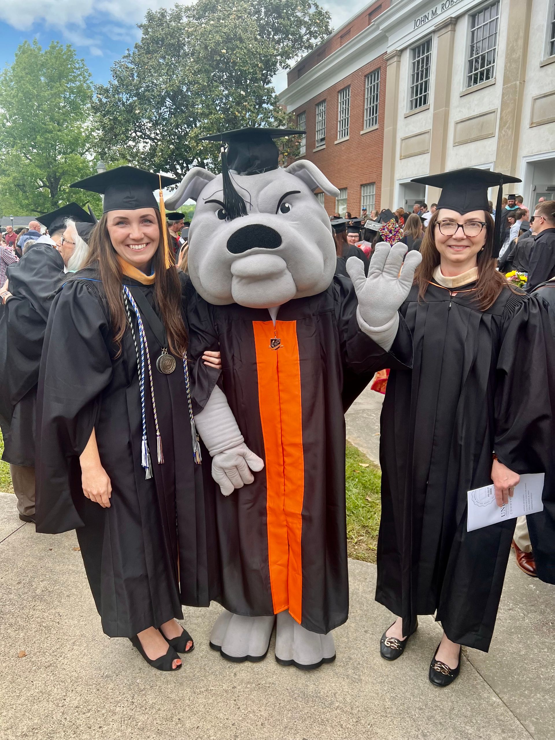 Lynn and Amanda with Union's mascot, Mack the Bulldog, all wearing caps and gowns on Union's campus, just after a commencement ceremony.