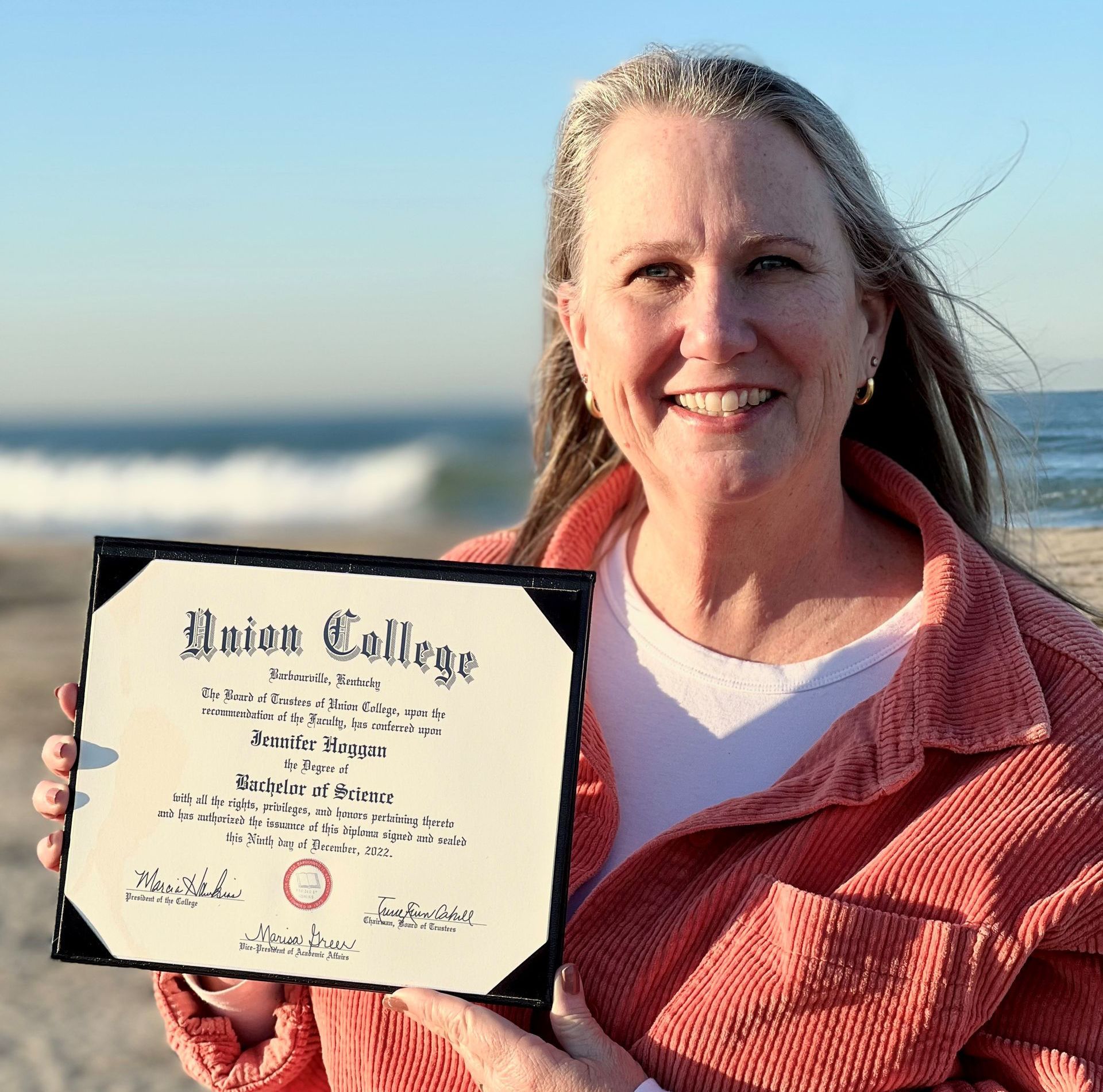 Jennifer Hoggan holding her Union College Diploma in front of the Pacific Ocean.