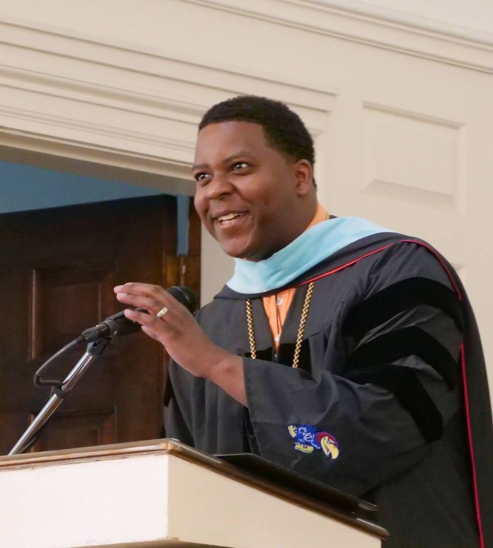 DJ Washington wearing academic regalia and talking into a microphone at a podium