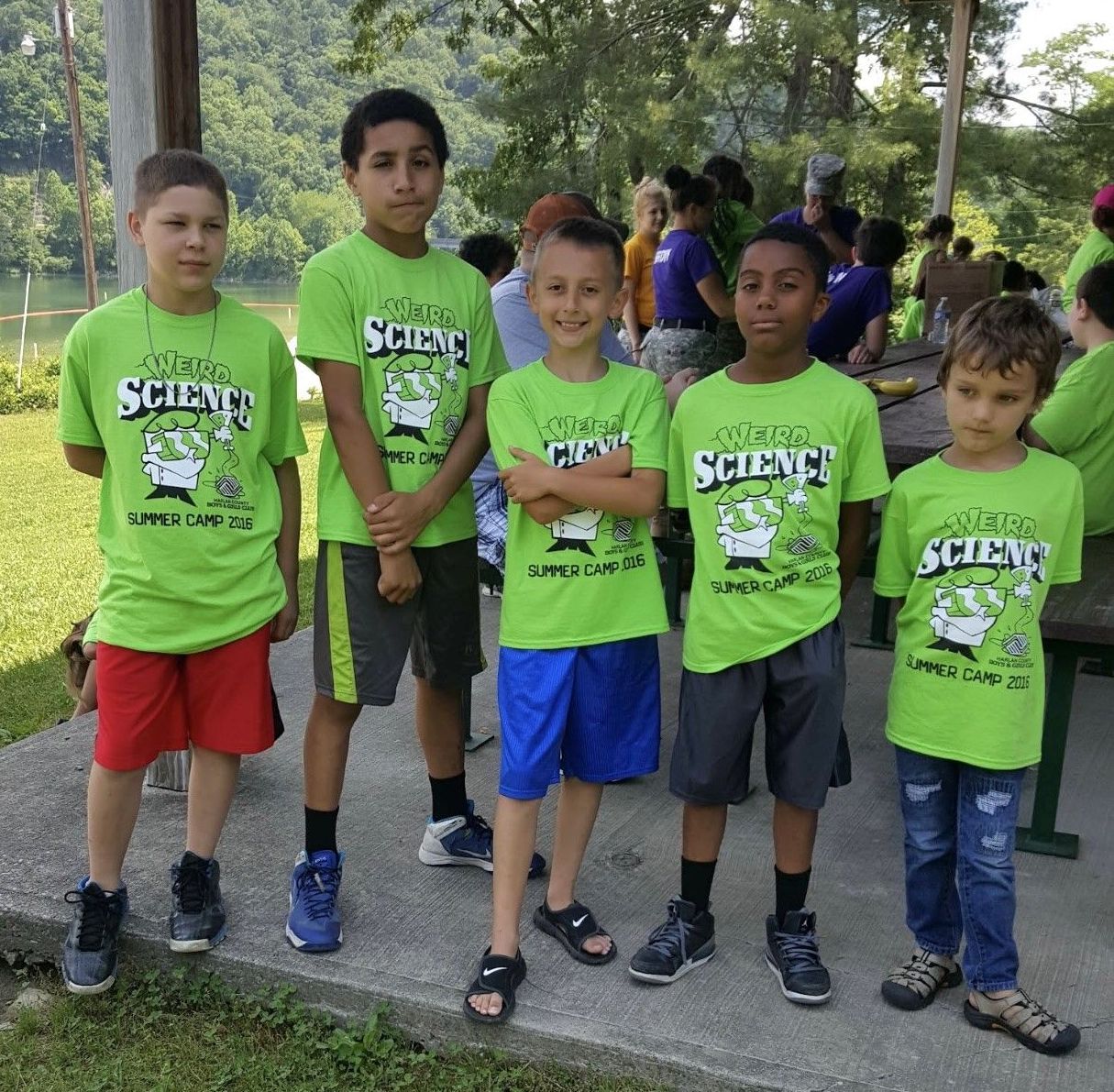 A young Kaleb, second from left, stands side-by-side with four other kids, all wearing lime green shirts that say Weird Science Summer Camp 2016. Lush green trees, a crowd of other children and adults, and picnic tables are visible in the background.
