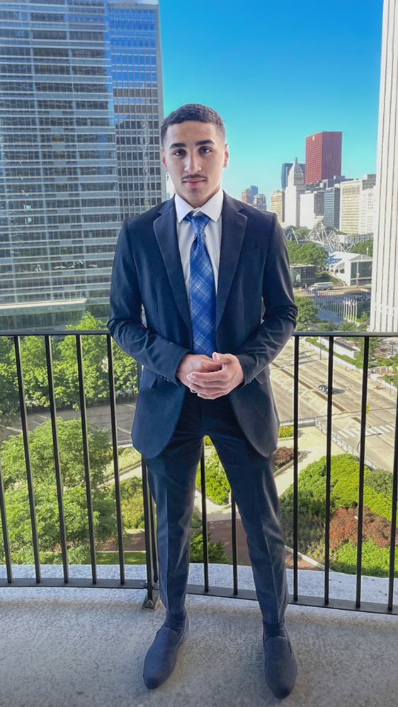 Kaleb McLendon stands on a balcony wearing a suit and tie. A Chicago cityscape with a few skyscrapers and roads below are visible in the background.