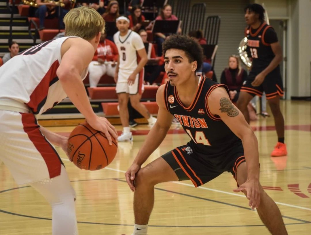 Kaleb, wearing a black and orange Union jersey with the number fourteen in white and orange on the front, faces off against an opponent in a red and white jersey on the basketball court. Other players and audience members in the stands are visible in the background.