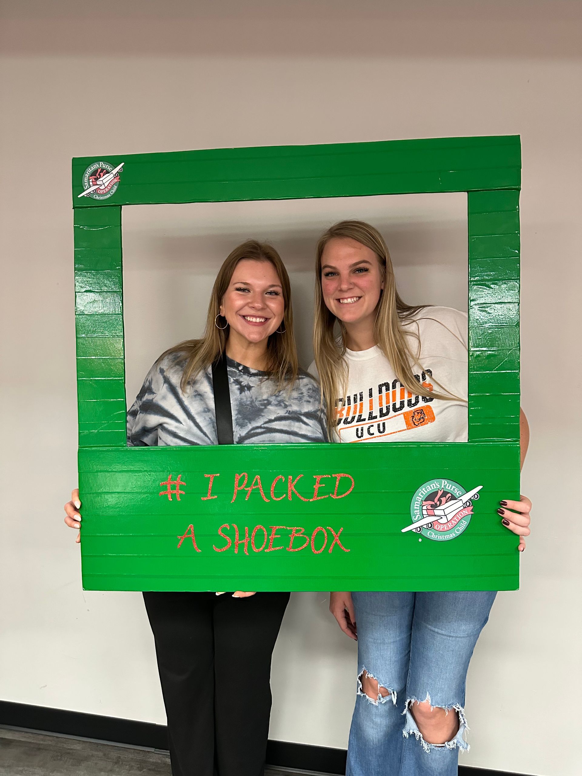 Kassidy Koogler and Allie Williams pose inside a green frame that says I packed a shoebox.