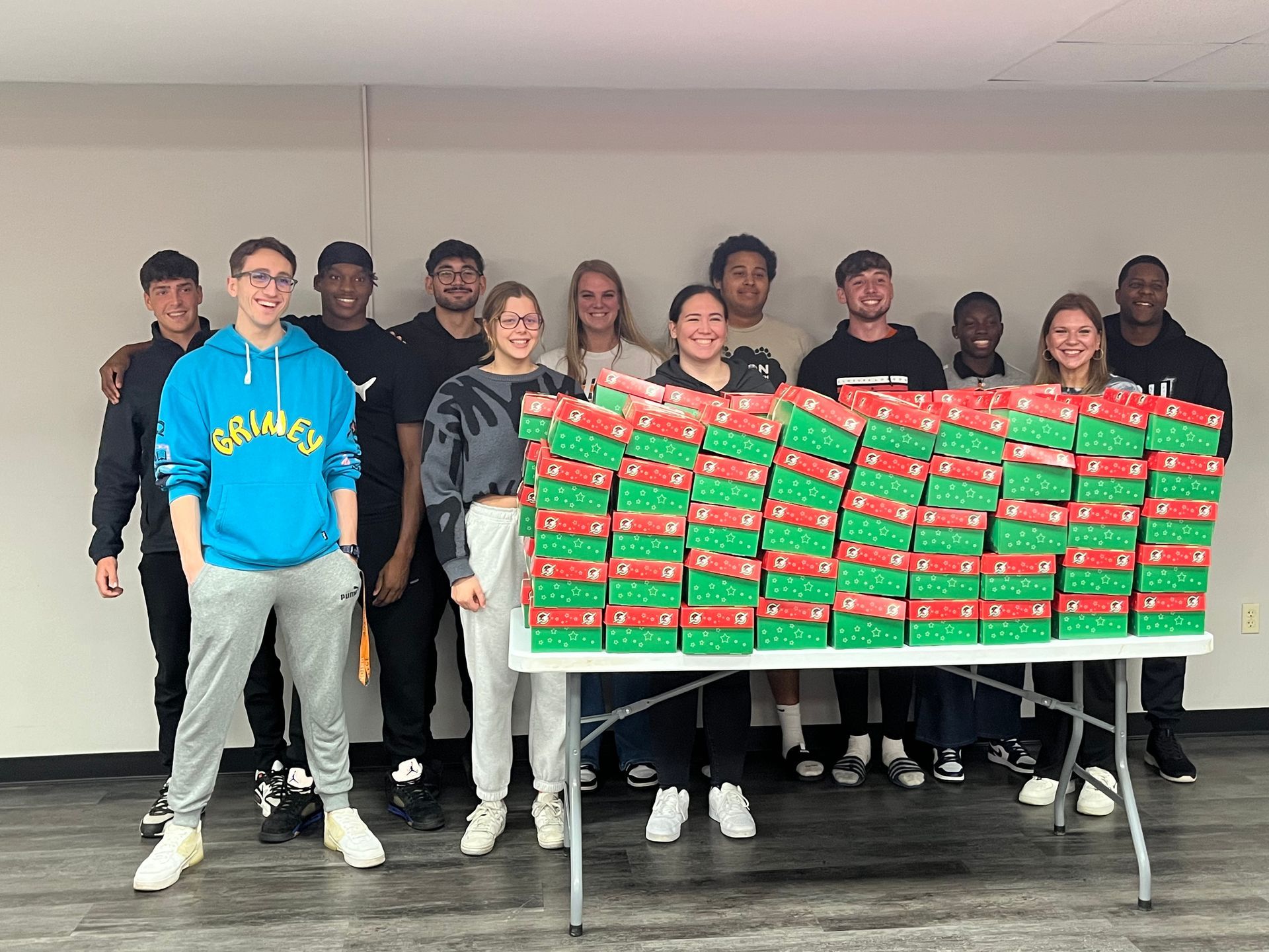 Smiling students, staff, and faculty members stand behind stacks of red and green shoeboxes.