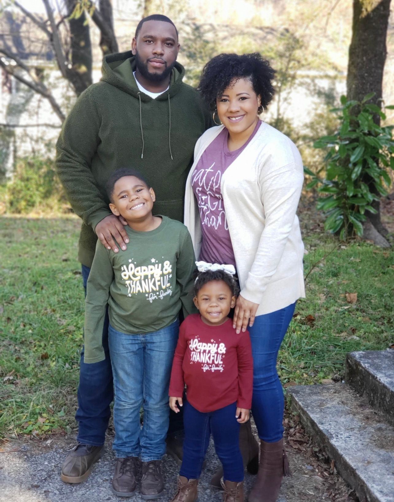 Harold and Kendra Calhoun stand with their young children in front of them, all smiling. Harold and Kendra have their arms around each other and their young children. In the background, a few trees and a white house are visible.