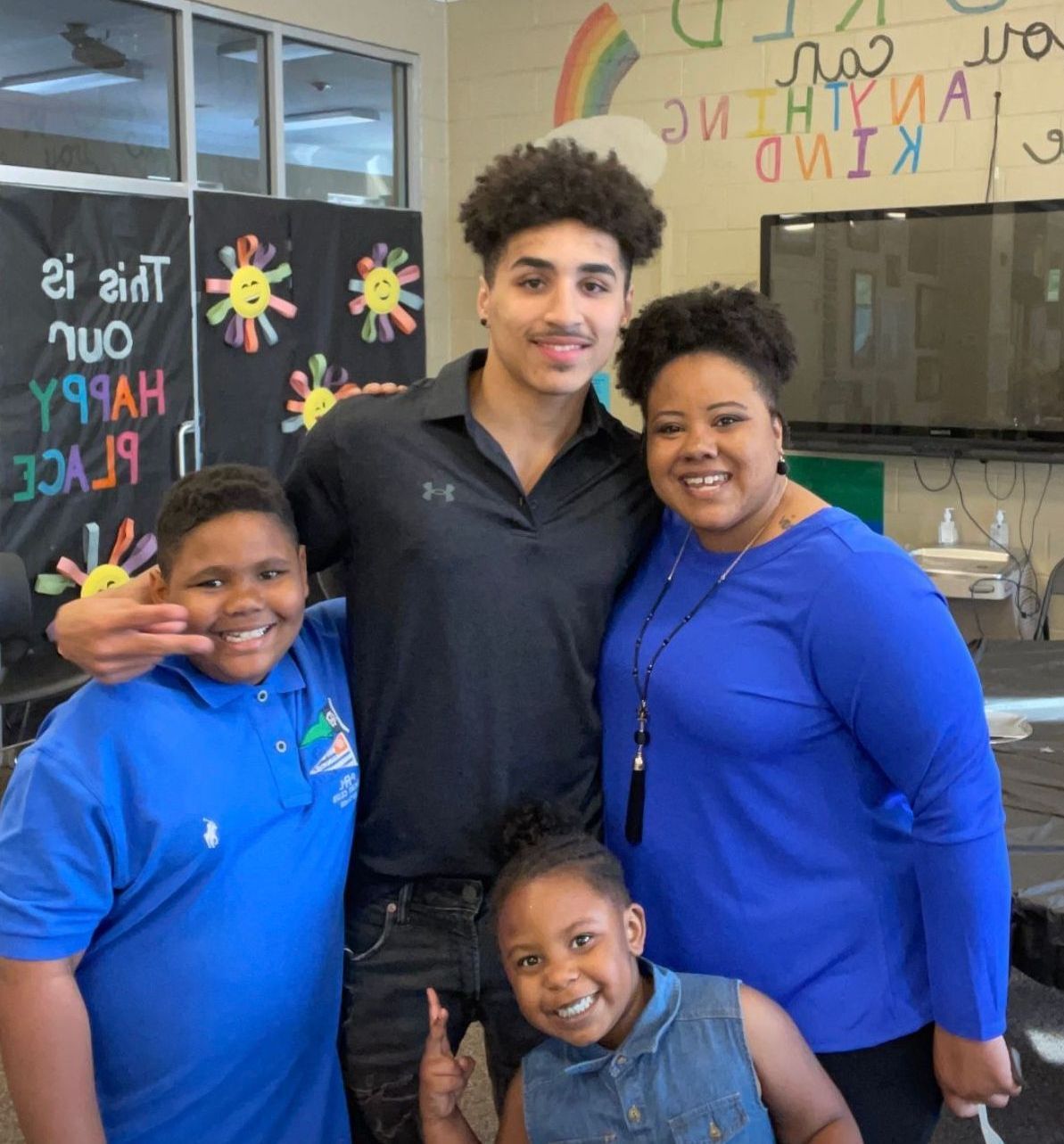 Kaleb McClendon poses with Kendra Calhoun and her two children in a classroom at the Boys & Girls Club. Kaleb has his arms around Kendra and her son. In the background, colorful writing can be seen on the walls alongside three rainbow sun crafts.