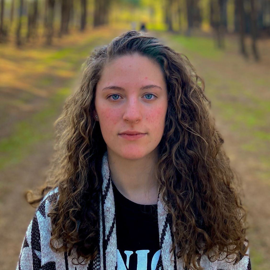 A photo of Union graduate student Mariane Tressoldi Moschetta wearing a blue and black patterned outfit. Behind her, a dirt road leading beyond some trees is visible.
