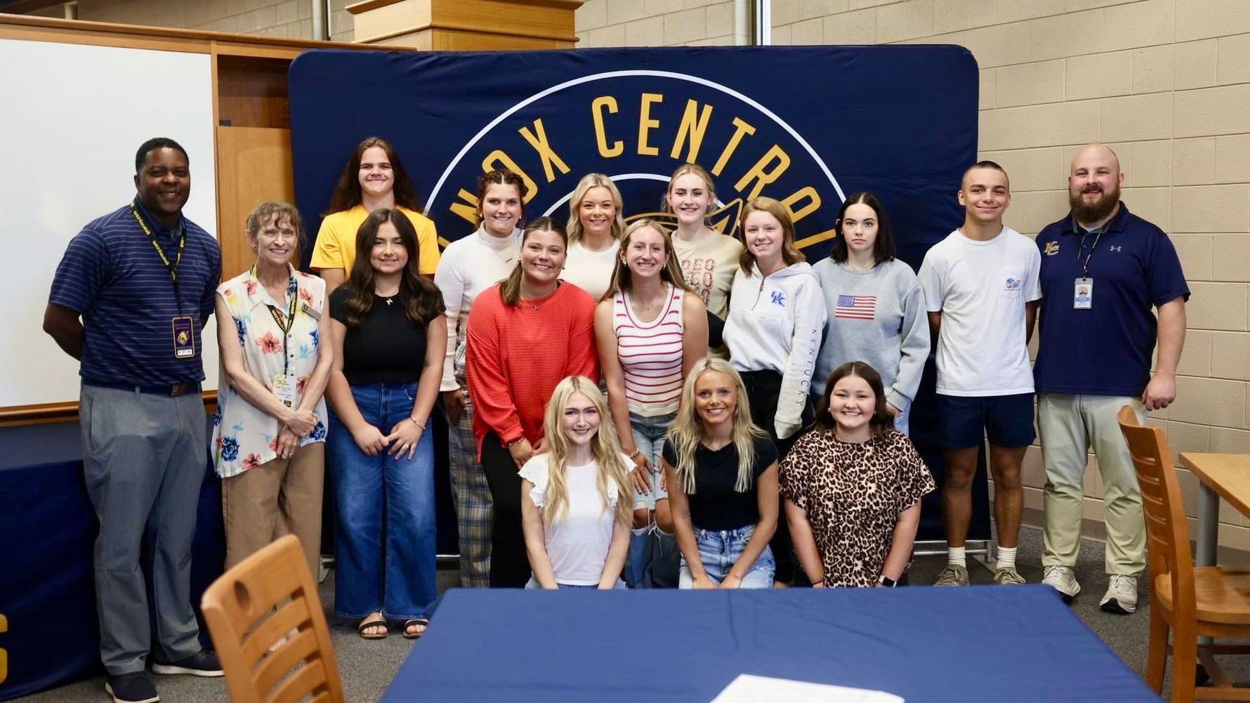 Knox Central students and adults pose in front of a dark blue school banner with yellow lettering. There is a whiteboard in the background and tables and chairs in the foreground.