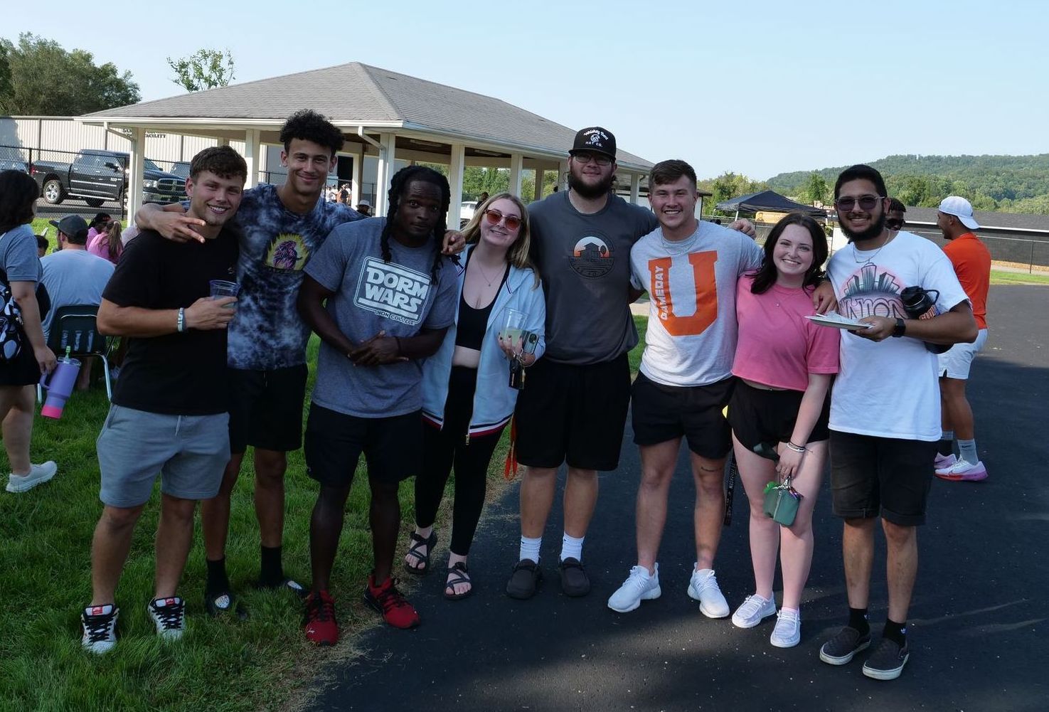 Eight students stand shoulder to shoulder on lush grass and a blacktop, smiling, some with their arms around each other's shoulders. Tyce is making a peace sign with two fingers, and some other students are holding plastic cups and a paper plate. Behind them, other students, a pavilion, and mountains are visible.