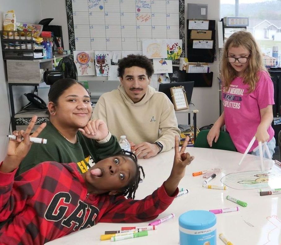 Kaleb McLendon and another club worker smile as they sit at a table with two girls in a Boys & Girls Club classroom. One child, to Kaleb's right, wears extended paper fingers, while the other leans on the table in front of them, sticking out her tongue and making peace signs with both hands. Art supplies are scattered across the table, and a desk and large calendar are visible in the background.