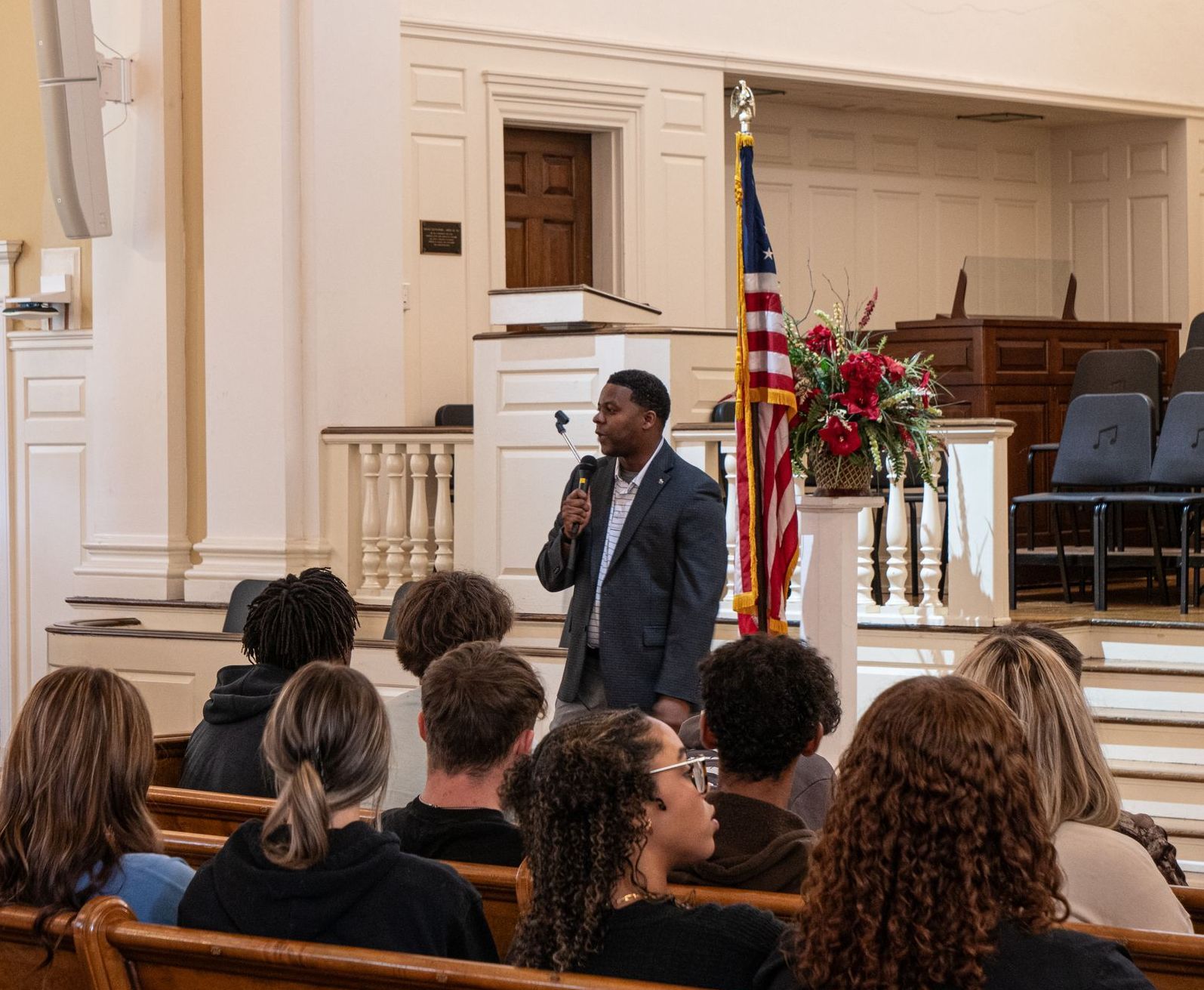 Washinton is standing in front of the podium, American flag, and flower arrangement in Cowan Chapel holding a microphone and wearing a suit jacket.