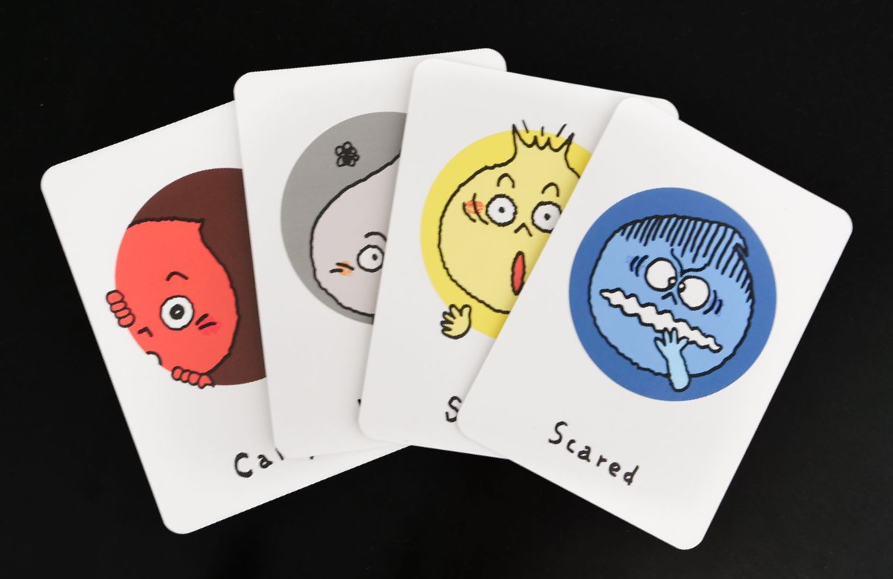Four musical expression flashcards are arranged against a black background, each showing a different expression. The card on the far right features a blue face with a fearful expression and the word Scared beneath it. To its left, a card with a yellow face shows a shocked expression. The remaining two cards, one with a gray face and the other with a red face, are mostly obscured by the first two cards.