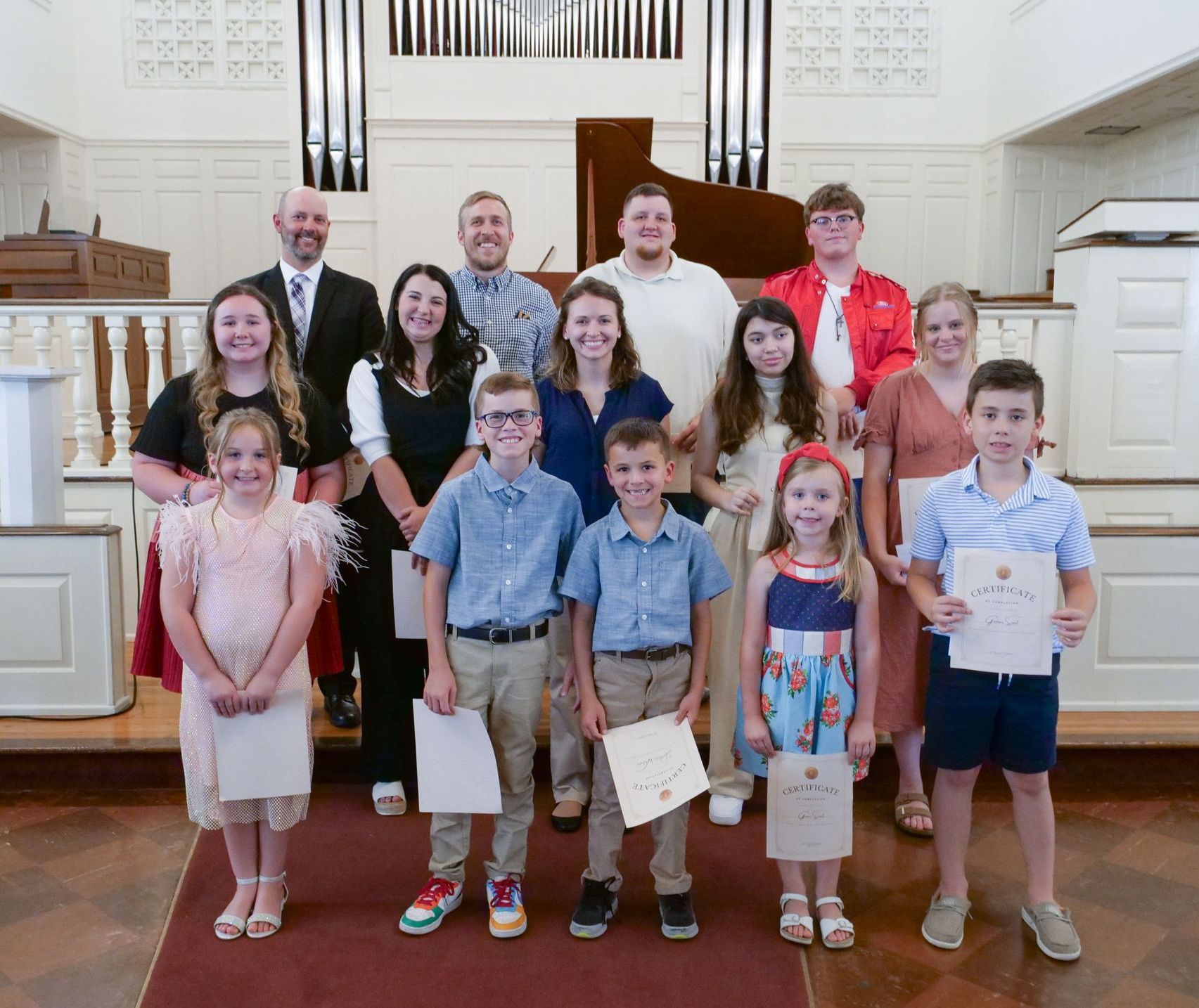A group of fourteen students, with adults in the back and kids in the front, stand together at the front center of the Conway Boatman Chapel holding their certificates of completion after their recital. They are dressed in formal or business casual attire, and most are smiling.