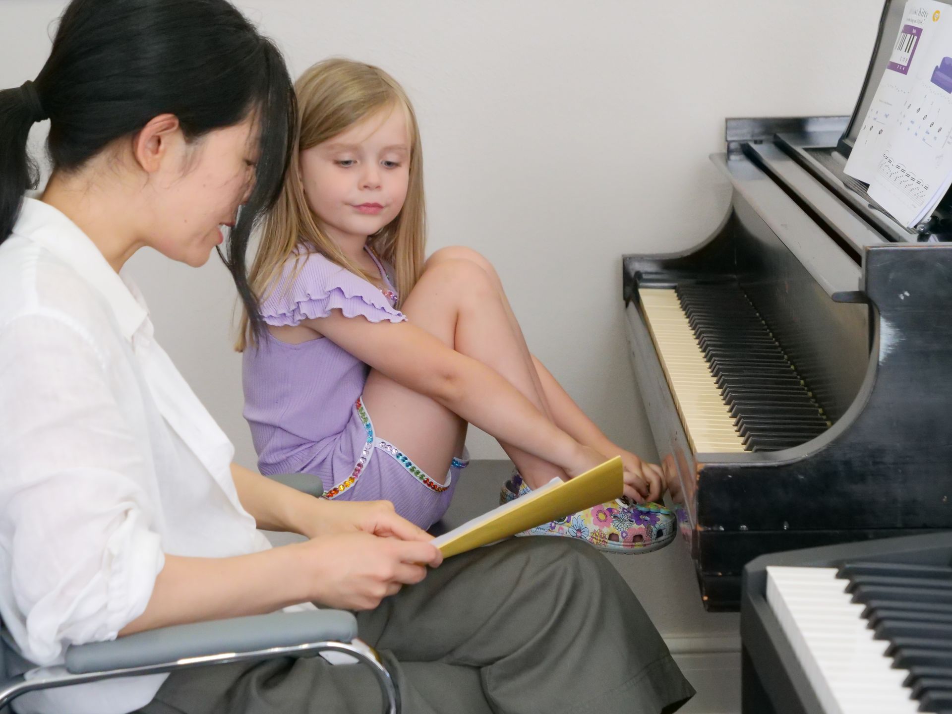 Dr. Fujimura and Grace Scent sit side-by-side at a grand piano and an electric piano. Grace sits in a huddled position with her knees up, looking toward the paper Fujimura is holding and pointing to. On the piano in front of Grace, additional sheets of music are visible.