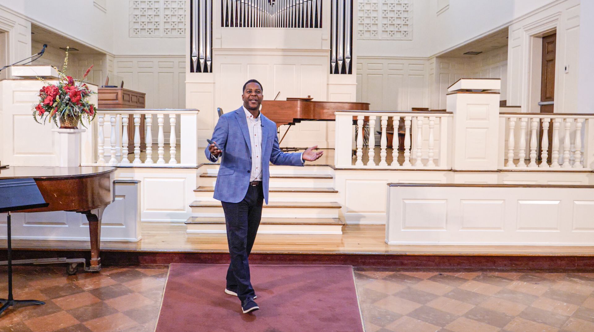 Dr. DJ Washington stands at the front center of the Conway Boatman Chapel, smiling with arms open wide, delivering his address to the audience. A microphone, lecterns, pianos, a flower arrangement, and organ pipes are visible in the background.