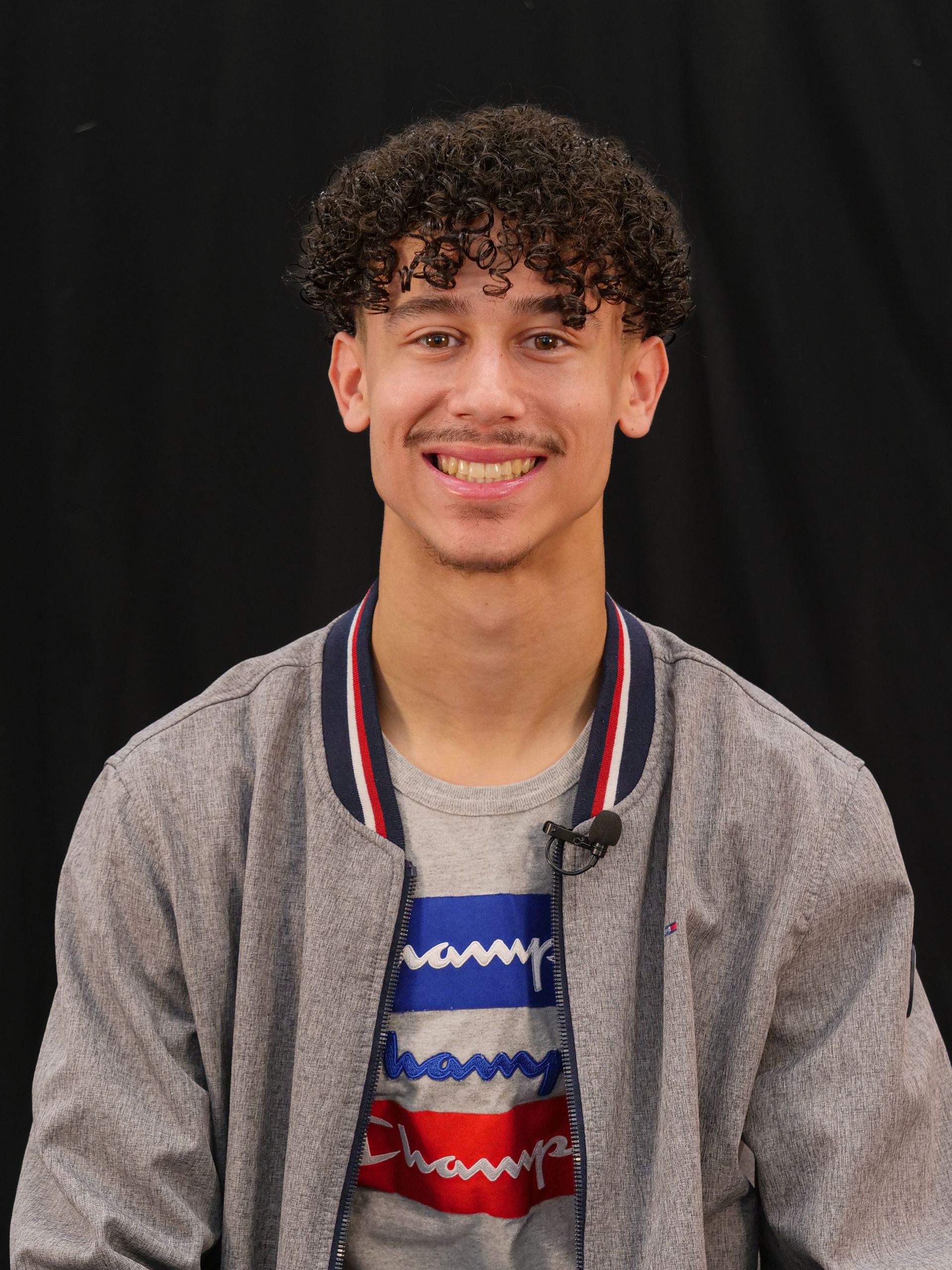 Headshot of Tyce: He is looking straight ahead, smiling, and wearing a t-shirt and lightweight jacket.