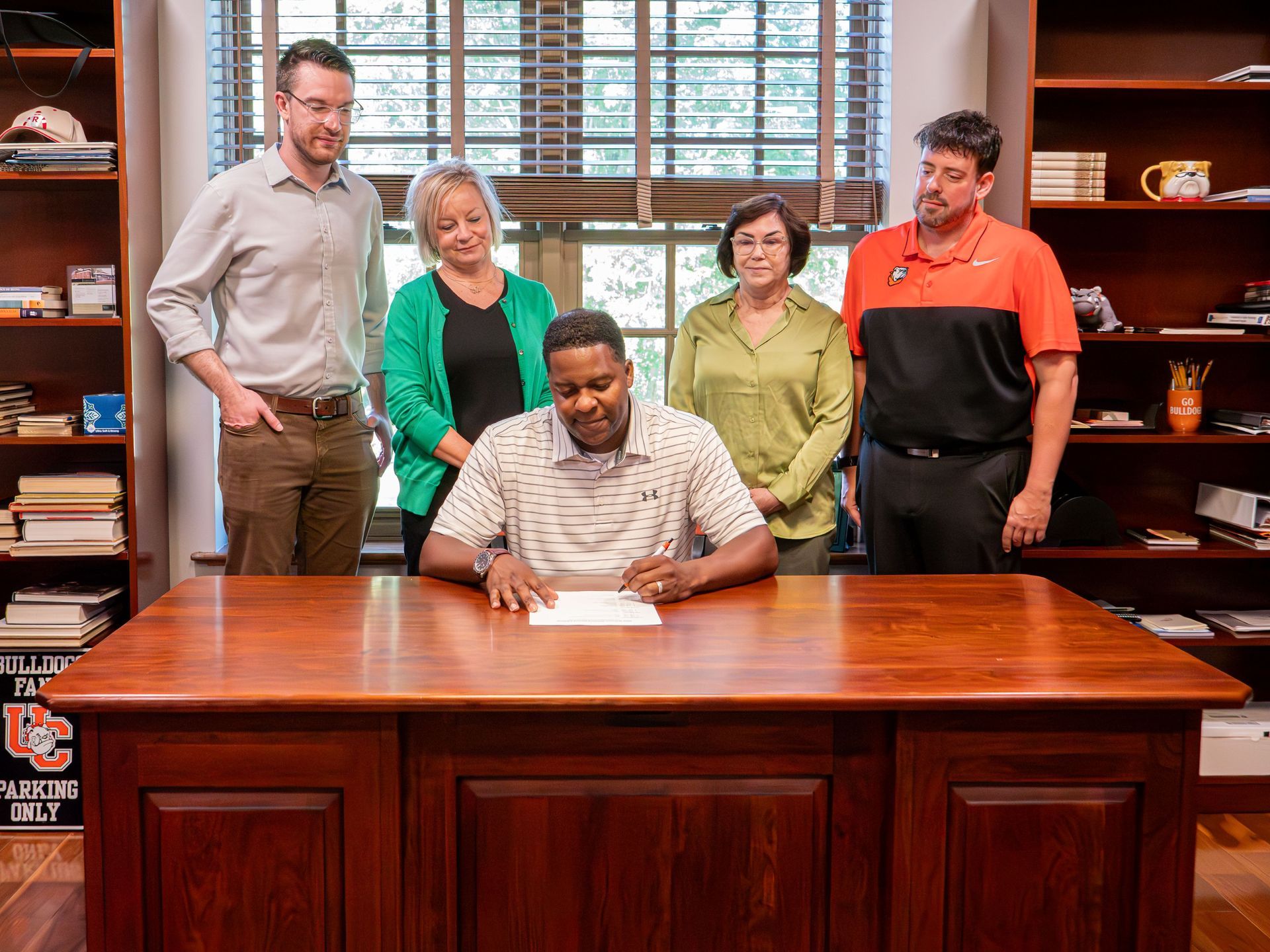 Dr . DJ Washington is seated at a wooden desk, signing a document. Standing behind him and gazing down at the document are Chris Gibson, Lisa Napier, Dr. Lori Henderson, and Dr. Ryan Winders.