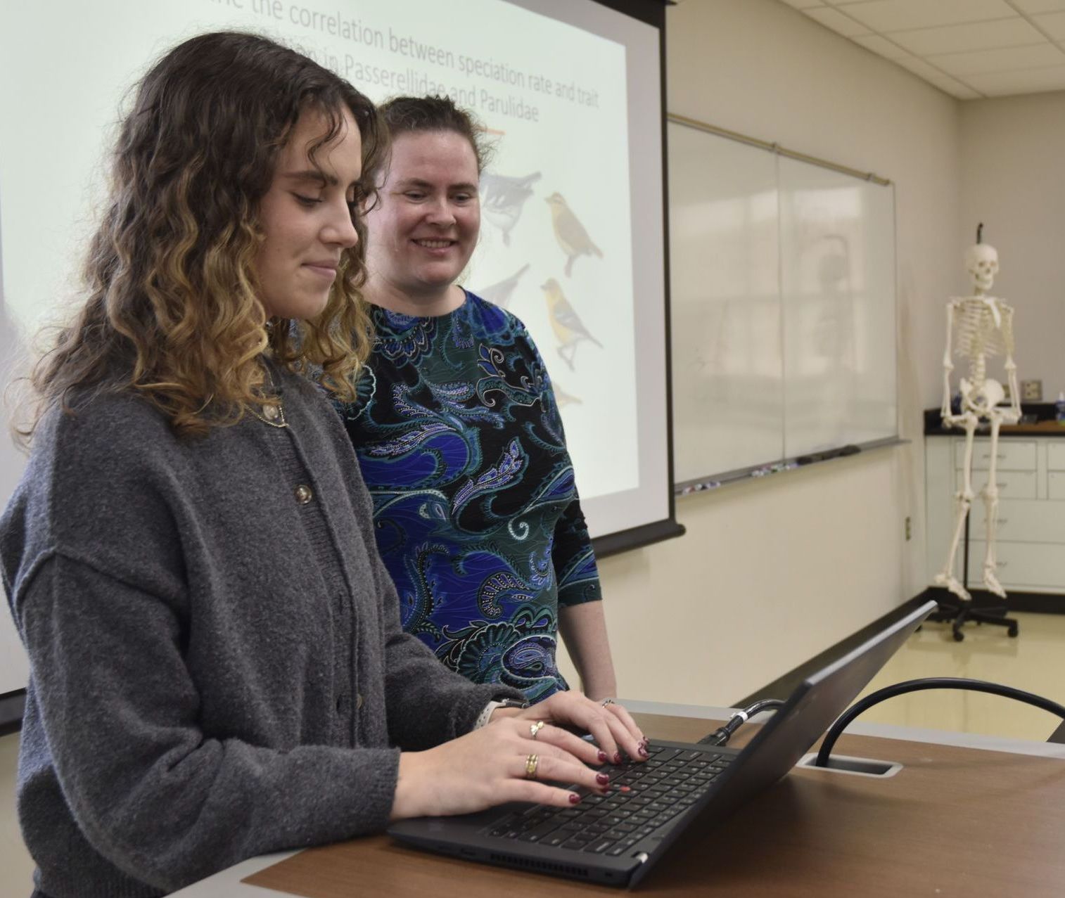 Lena typing on her laptop with Megna looking on supportively behind her.