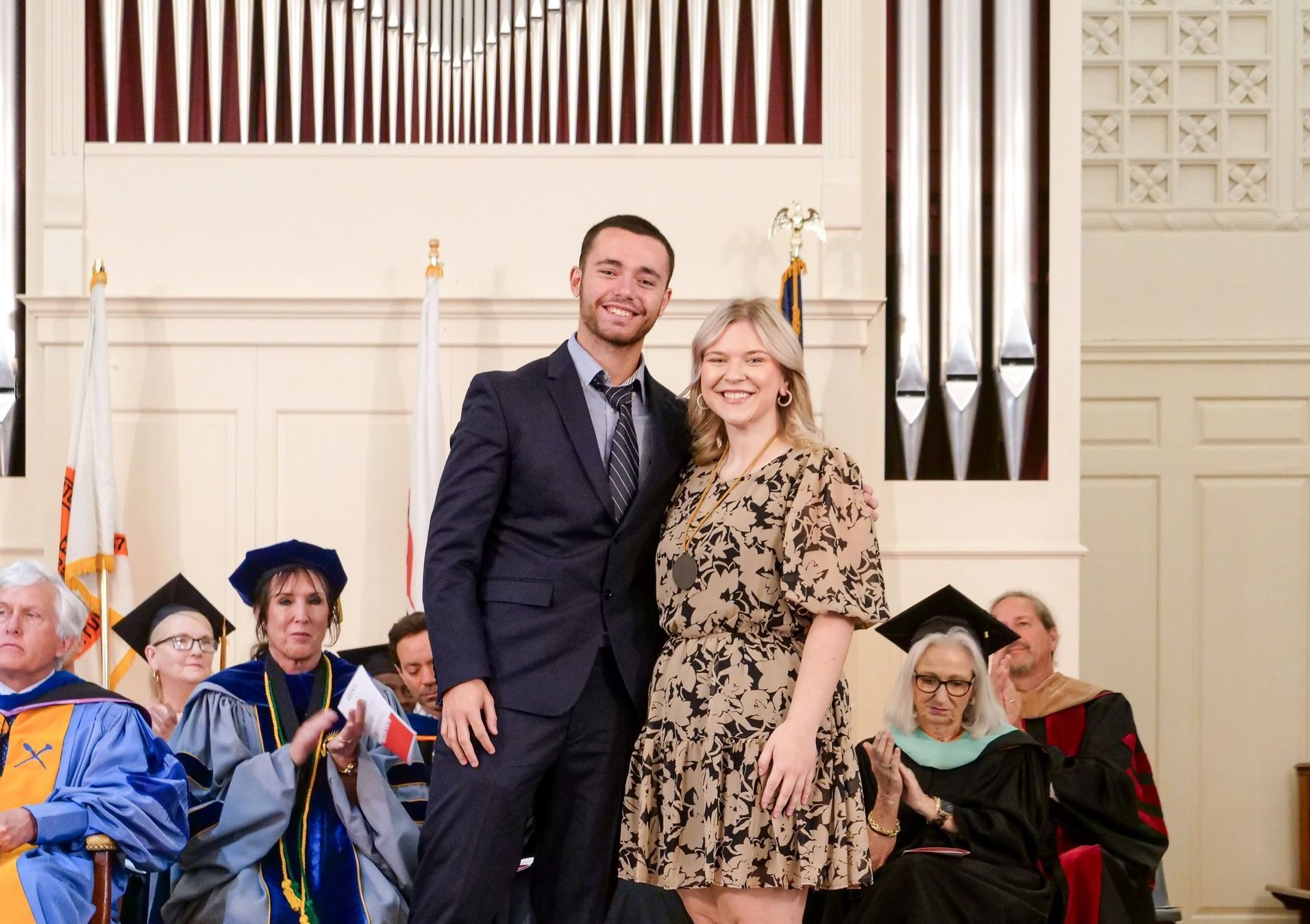 Carlos Sainz Barroso poses with his arm around Kassidy Koogler at the front of the chapel. Both are smiling, and Carlos is wearing a black suit while Kassidy is wearing a black and yellow dress. Behind them, seated faculty members wearing academic regalia and three flags are visible.