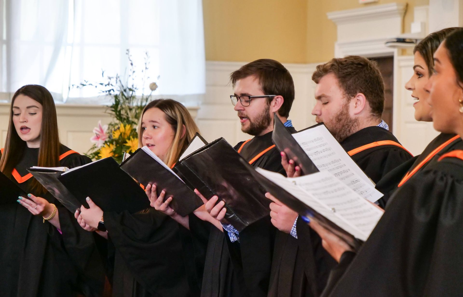 Six Union Harmony members wearing orange and black choir robes sing in the chapel while holding song sheets in black folders. Behind them, an arrangement of pink and yellow flowers is visible.
