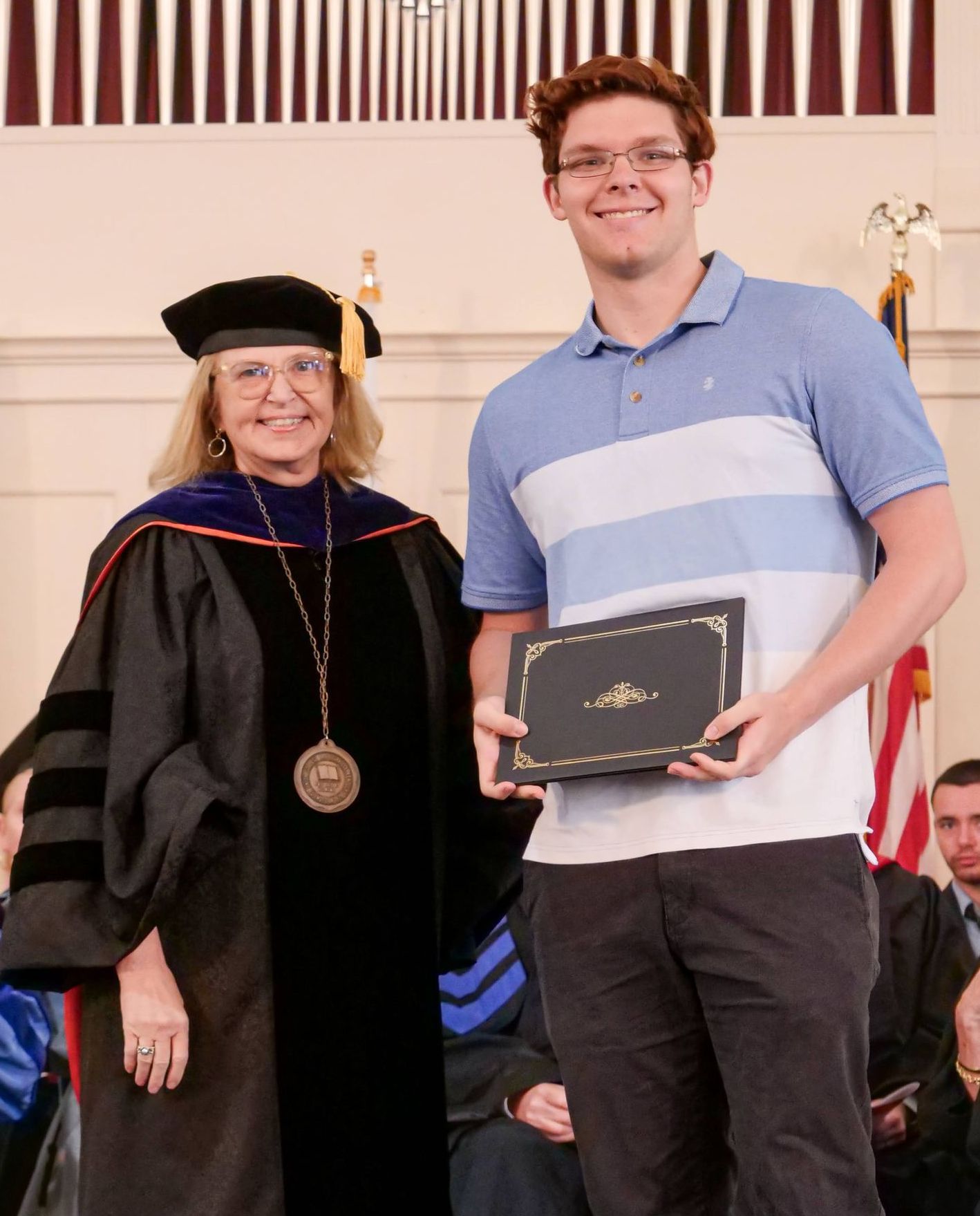 In the chapel, Webb stands side-by-side with Hawkins while holding his President's award, which is in a black certificate holder with gold trim. Both are smiling, and Hawkins wears her academic regalia while Webb wears a blue and white striped shirt with black pants.