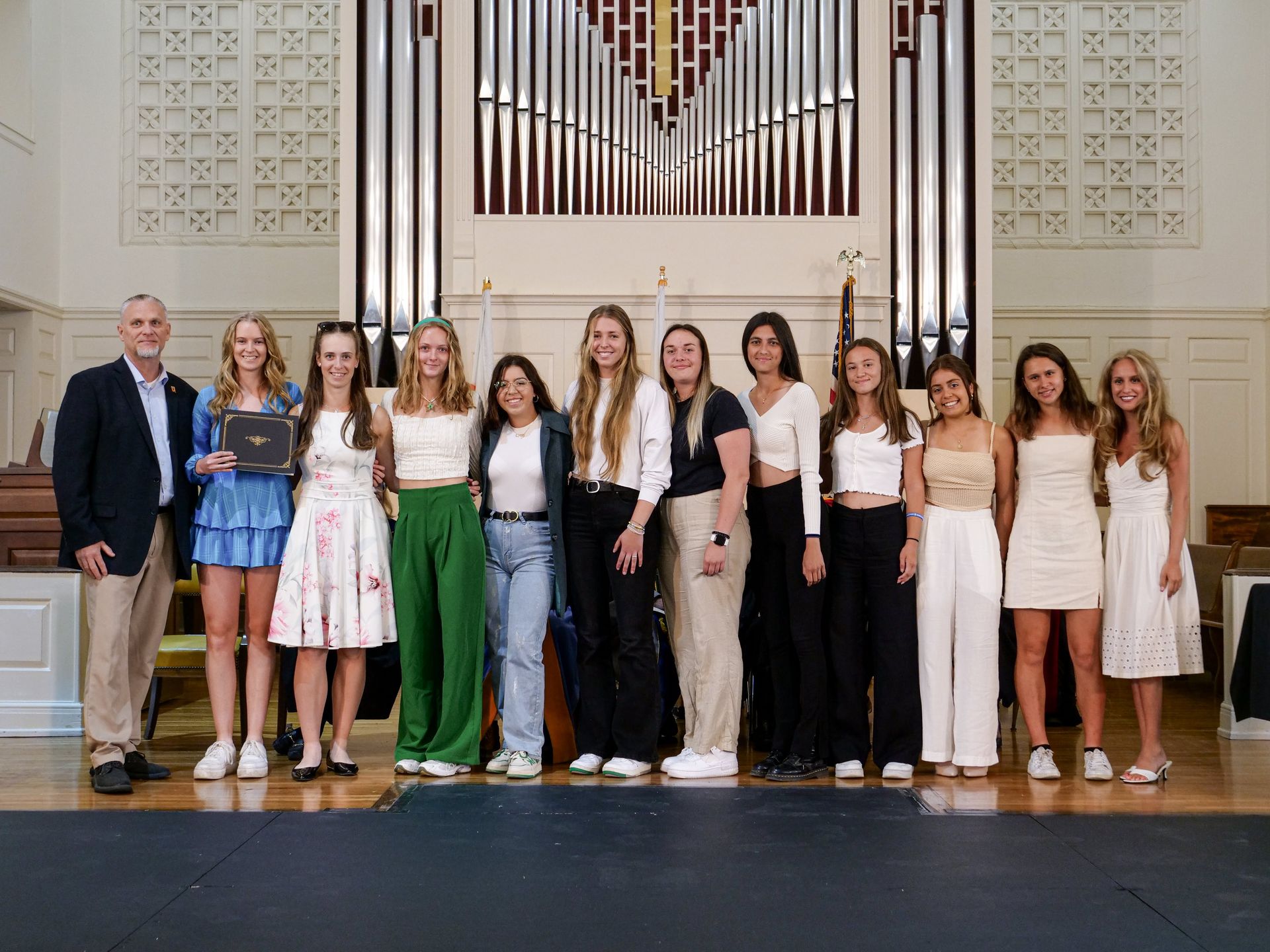 Eleven members of the Women's Tennis team in formal attire stand side-by-side at the front of the chapel with Athletic Director Tim Curry. One team member wearing a bright blue dress holds the team's award which is in black certificate holder with gold trim.