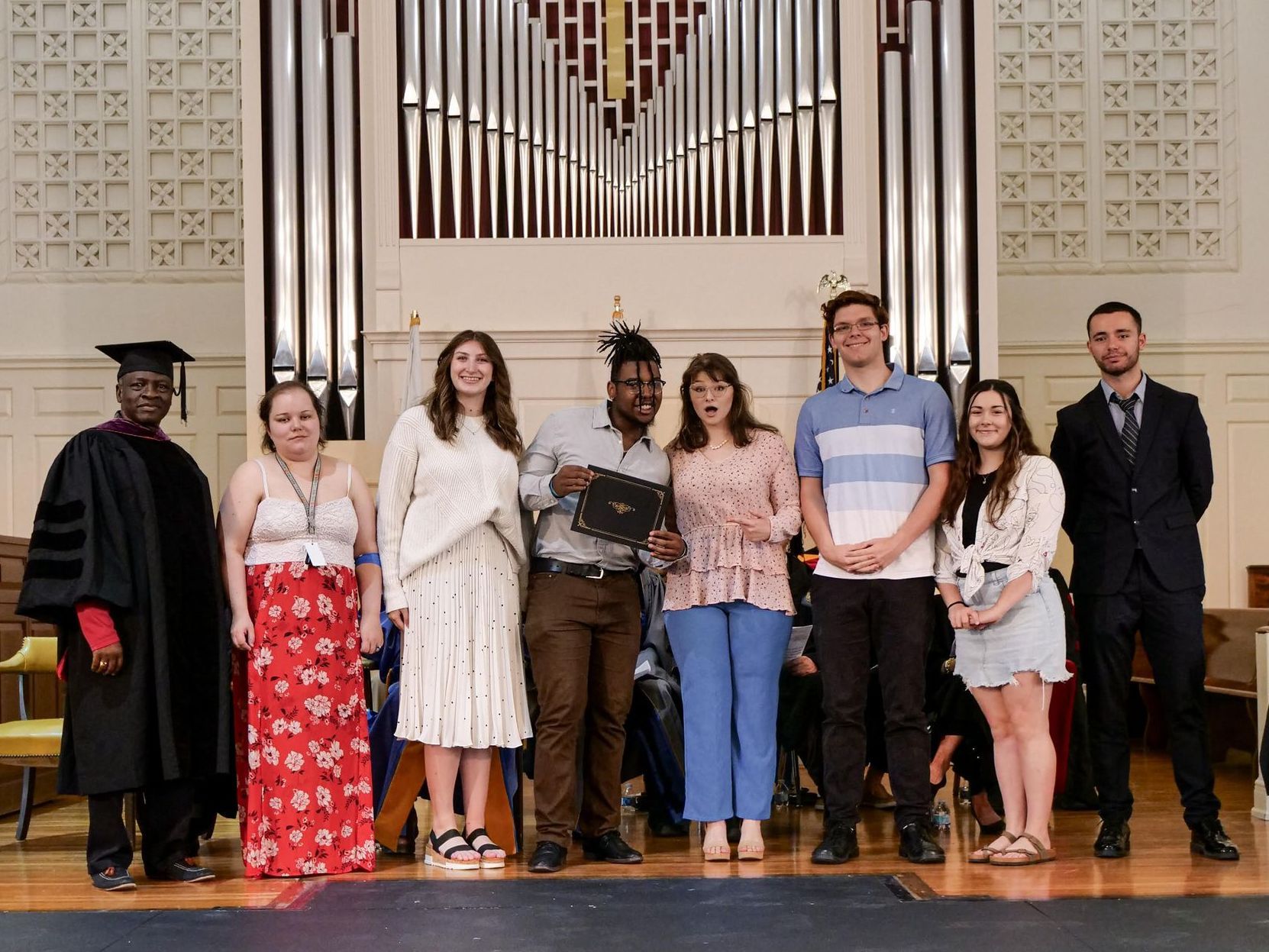 Seven Justice Leaders in formal and business casual attire stand side-by-side at the front of the chapel with Dr. Fidelis Achenjang. In the center, Malik McCalpine holds the award and leans into Maggie Heinrich, who is pointing to it with a mock shocked expression on her face.