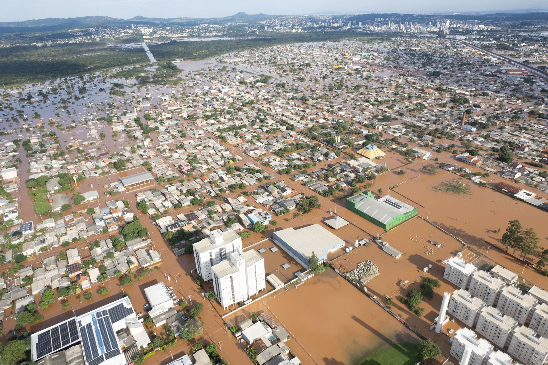 From a view above the Rio Grande do Sul cities of Novo Hamburgo and São Leopoldo, flooding on a wide scale is visible. Several buildings that look like apartments, offices, restaurants, and houses are covered in water. Additionally, many yards and roads are submerged. The upper portions of many buildings and the tops of many trees stand visible among the flooded area.