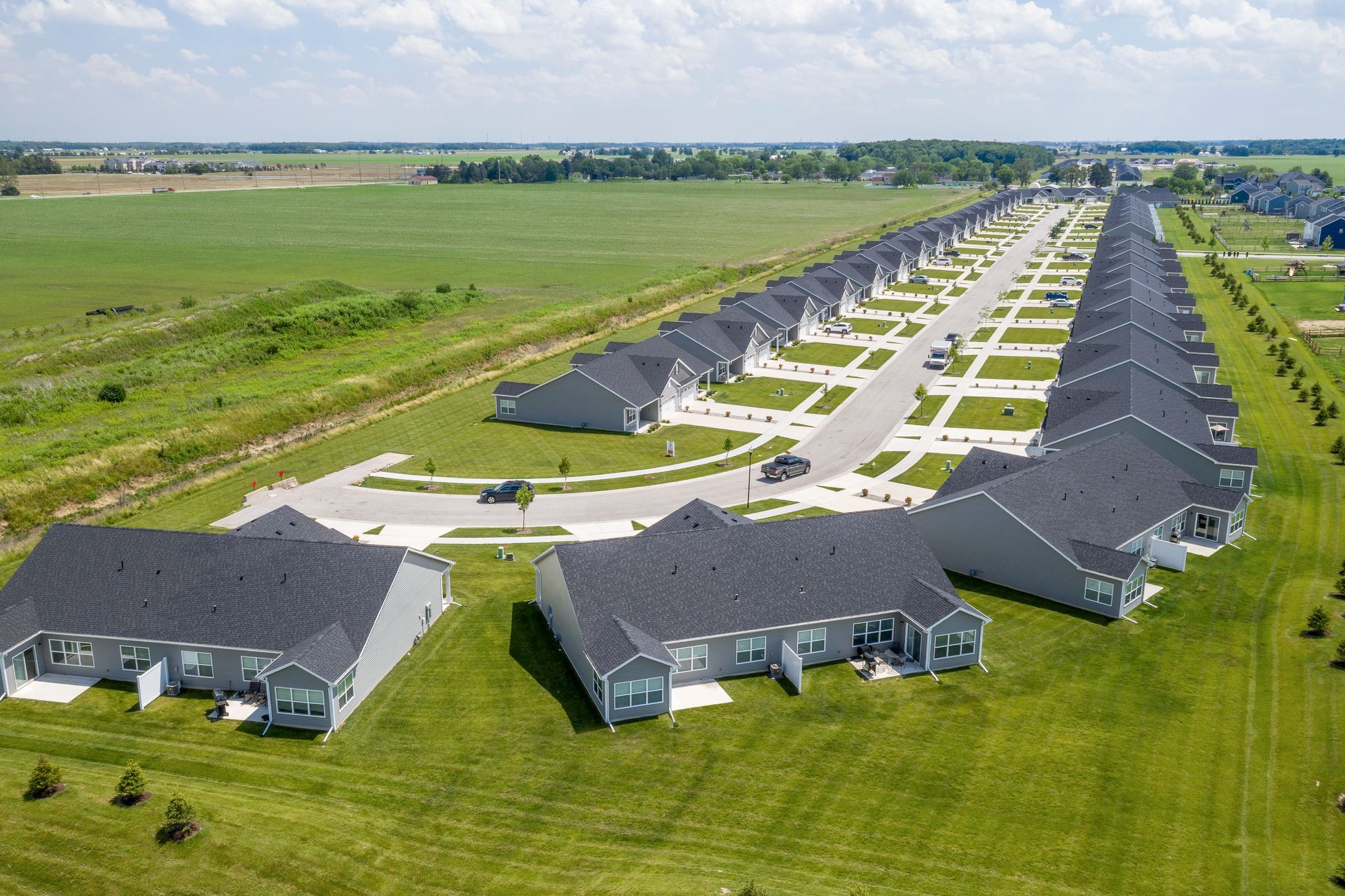 Aerial view of a long row of gray houses with dark roofs along a street, set in a grassy field.