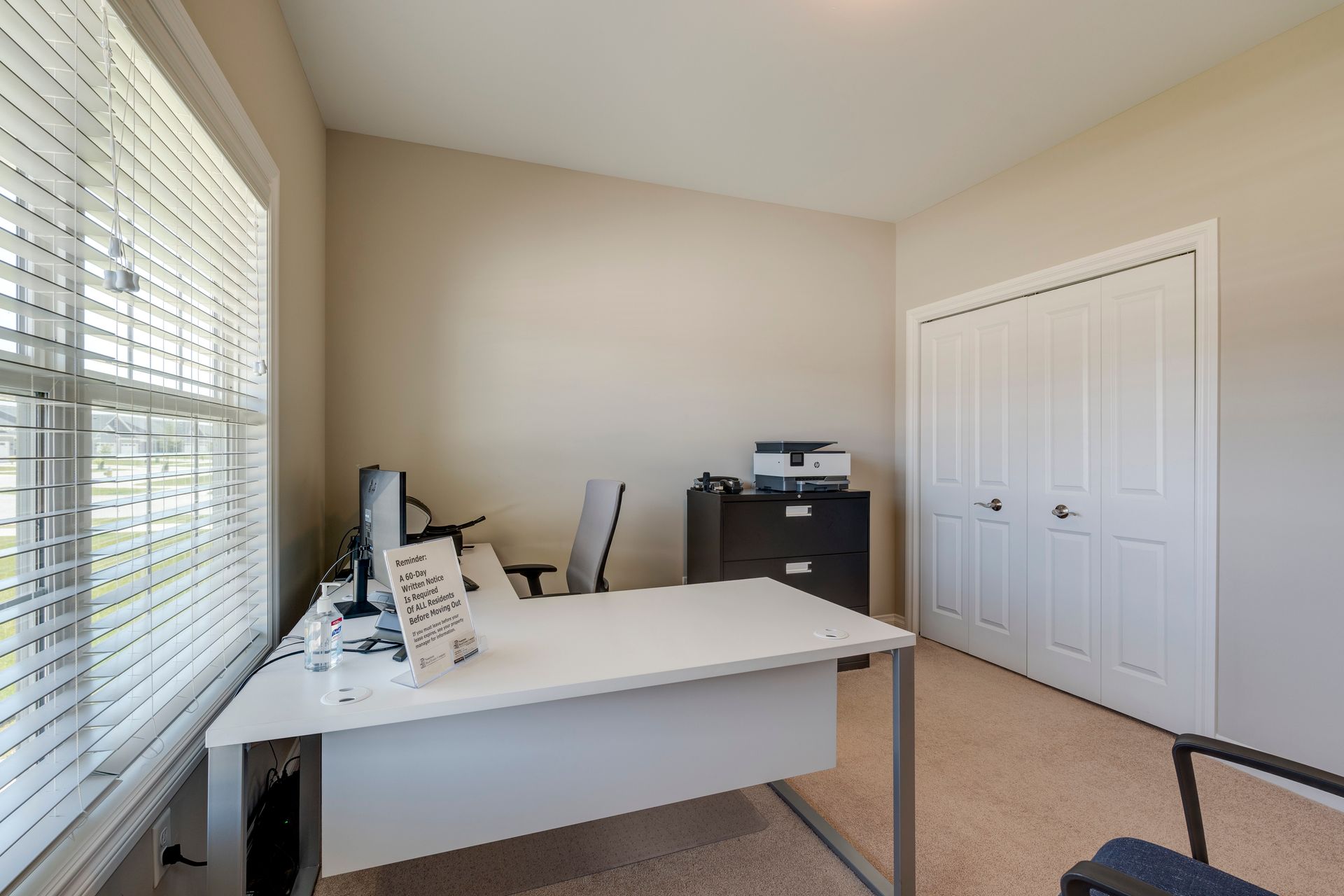 Office with a white desk, closed white closet, window with blinds, and file cabinet.