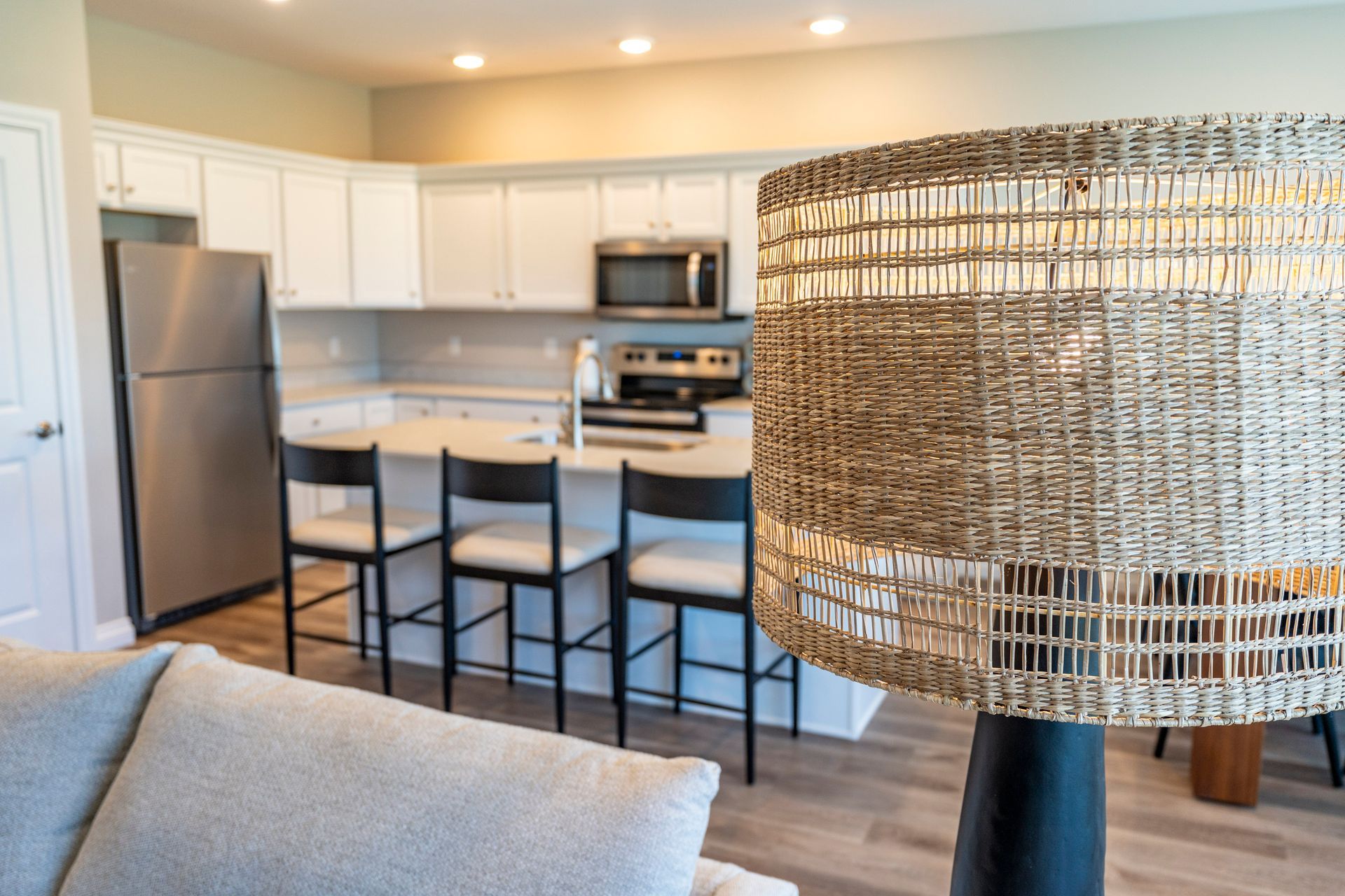 Kitchen with white cabinets, stainless steel appliances, and island with bar stools. Woven lamp in the foreground.