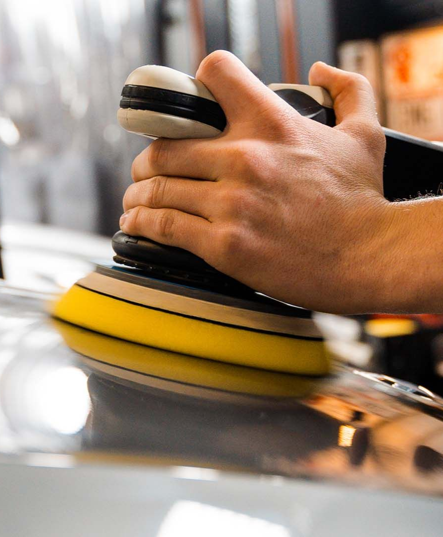 Hand using an electric polisher with a yellow buffing pad on a vehicle's surface.