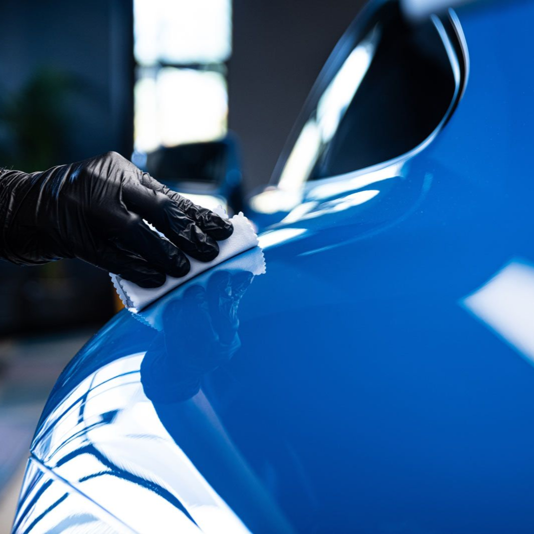 Gloved hand polishing a shiny blue car's curved surface with a white cloth.