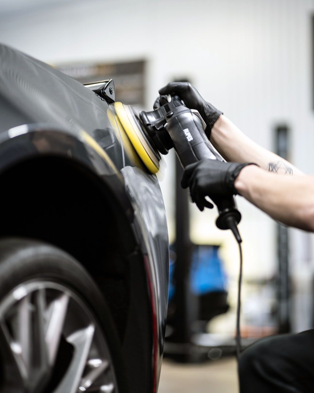 Person wearing black gloves polishing a black car with a buffer in a garage.