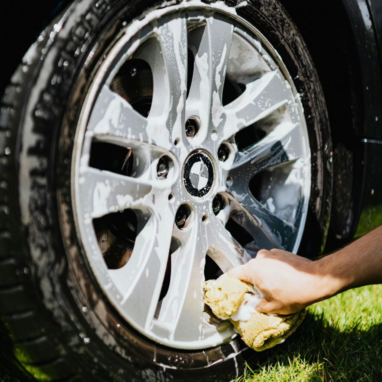 Person washing a silver car rim with a sponge, suds visible, outdoors on grass.