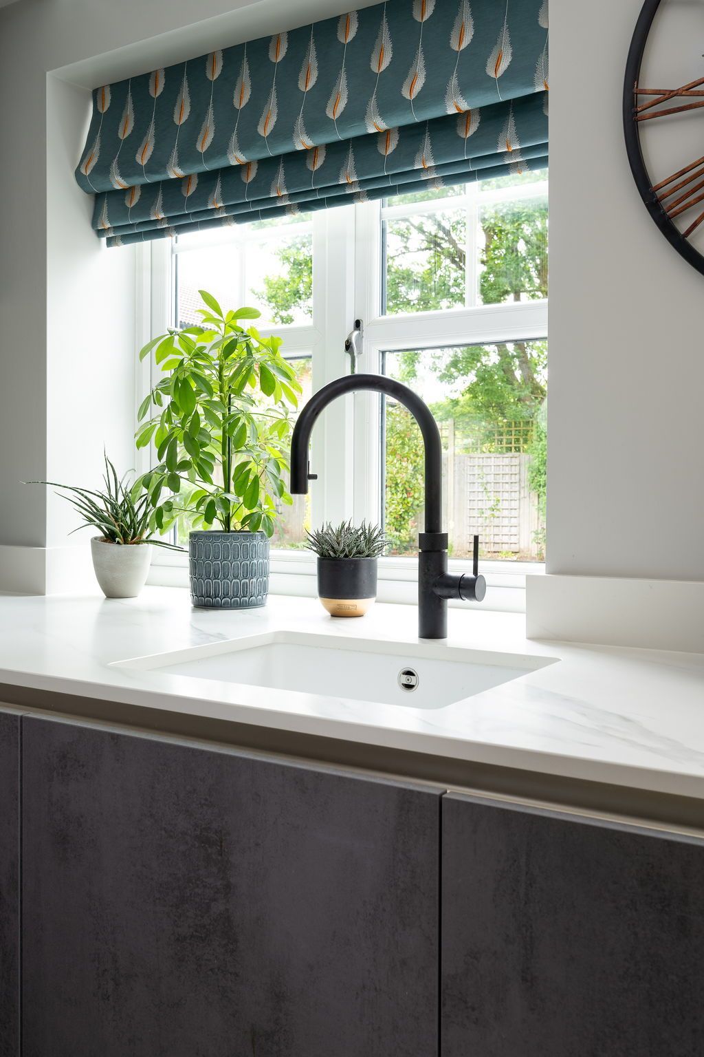 Kitchen sink with a black faucet in front of a window with plants and patterned shades. Gray cabinets.