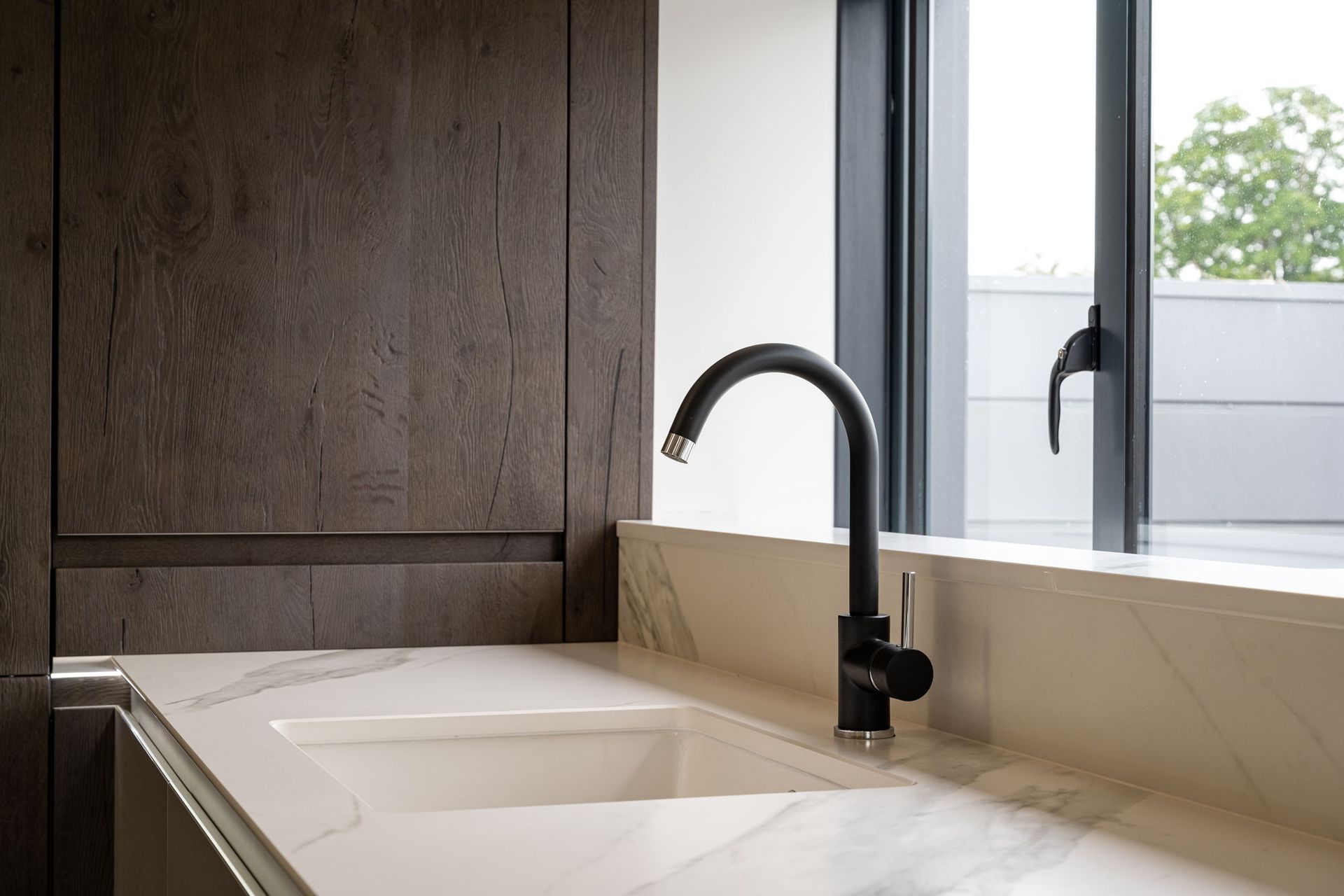 Kitchen sink with black faucet, white countertop, dark wood cabinets, and window with outdoor view.