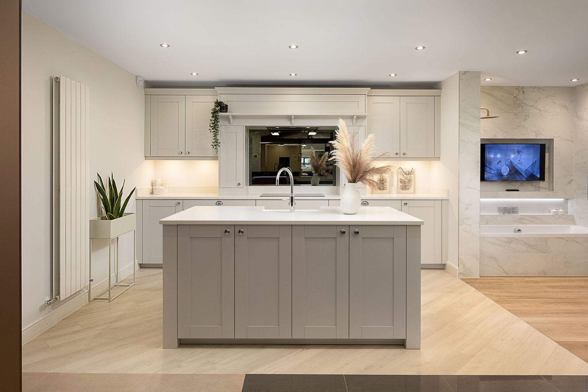 Modern kitchen with island, pale gray cabinets, and light wood flooring. A television is built into the wall.