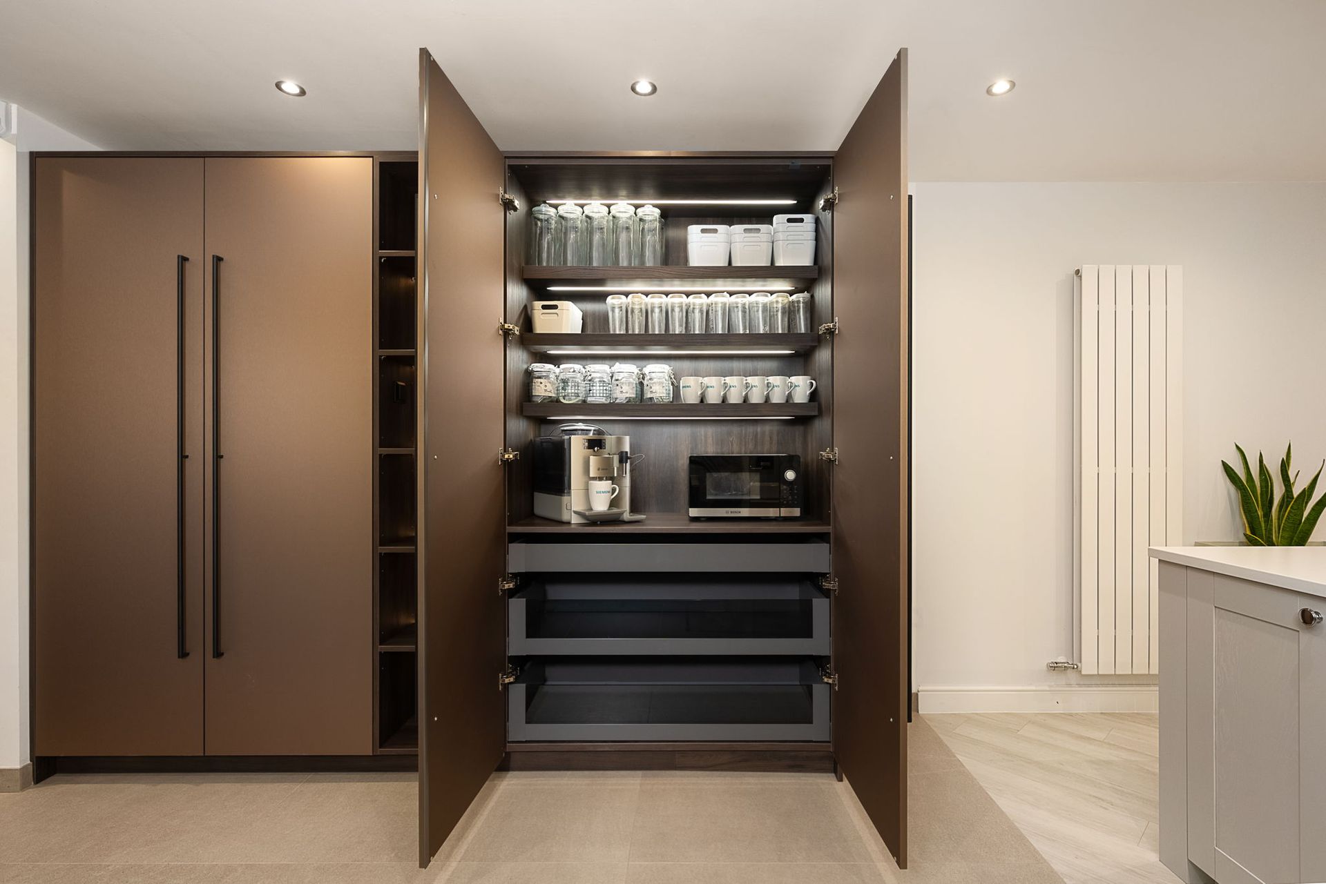 Kitchen pantry cabinet with open doors, shelves with coffee maker, microwave, and storage. Brown cabinet, white wall.