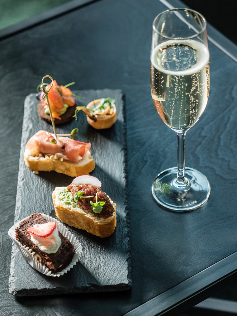 Champagne flute beside a slate tray with assorted hors d'oeuvres: toast, pastry, and a small quiche.