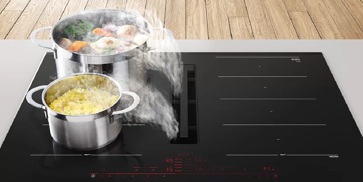 Black stovetop with pots boiling vegetables and grain, steam rising.